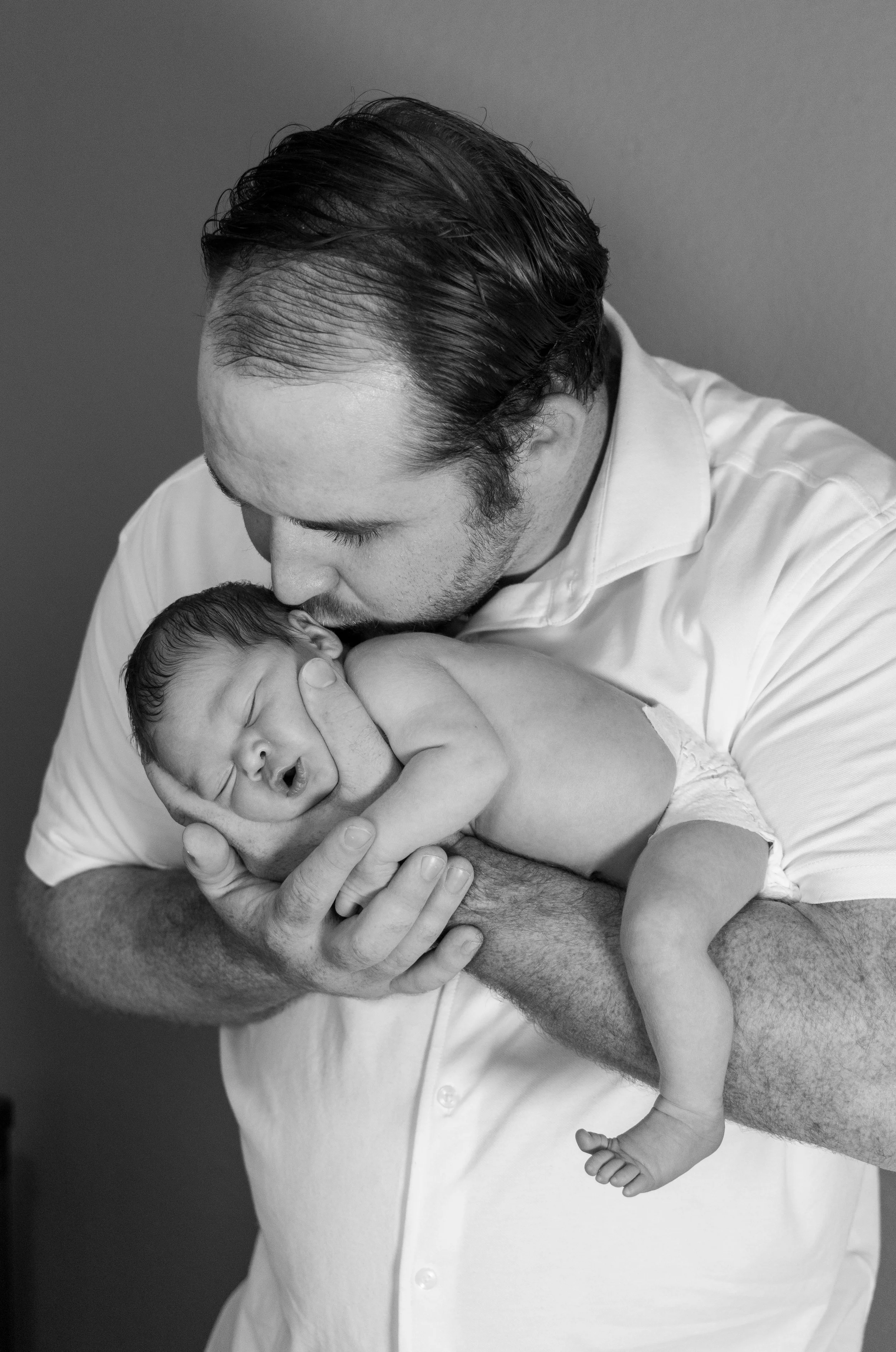 A man holding a newborn baby close to his chest, with the man's face leaning down to kiss the top of the baby's head. The image is in black and white.