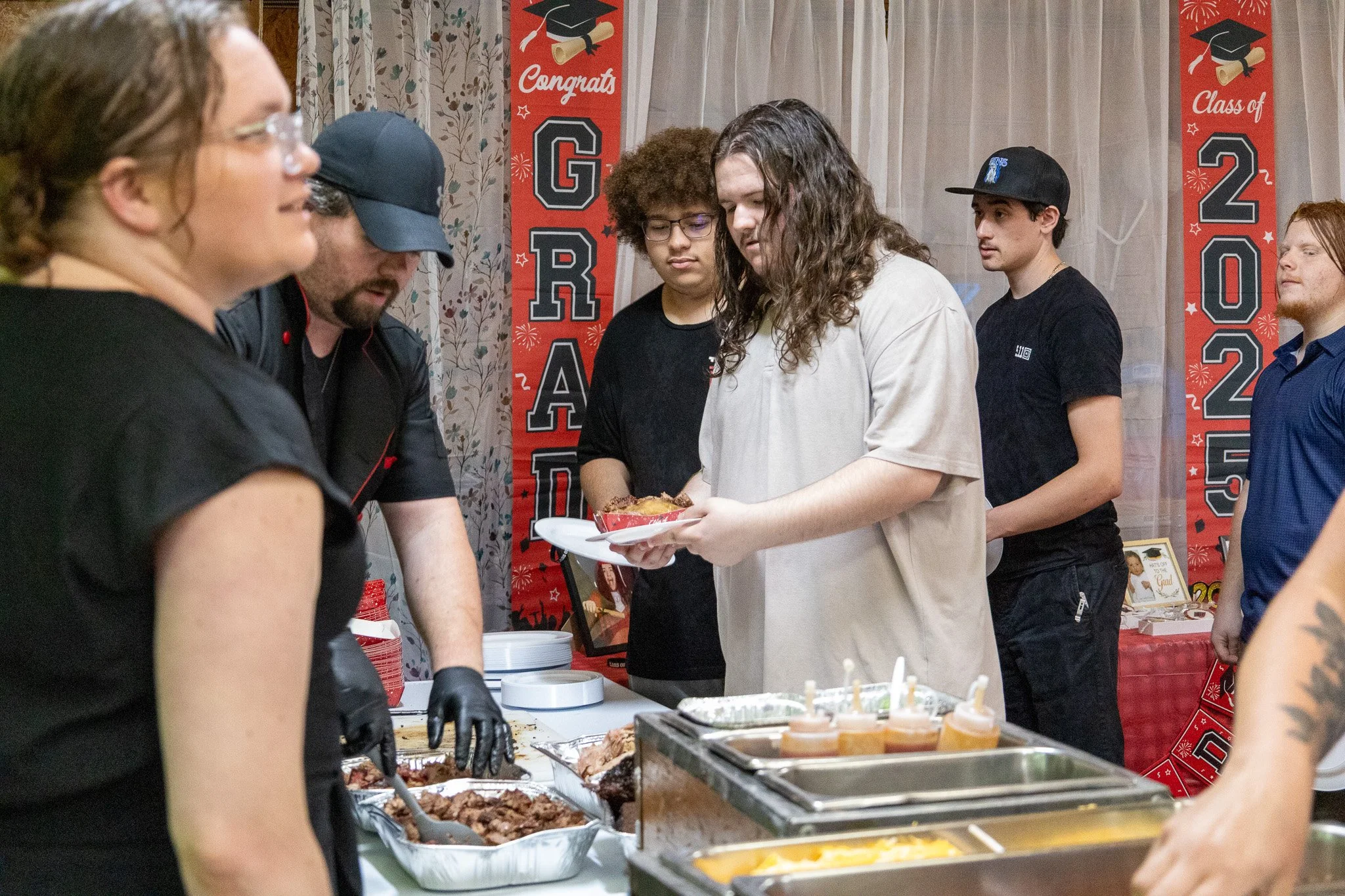 Students and staff serving food at a graduation celebration with red banners and table of food.