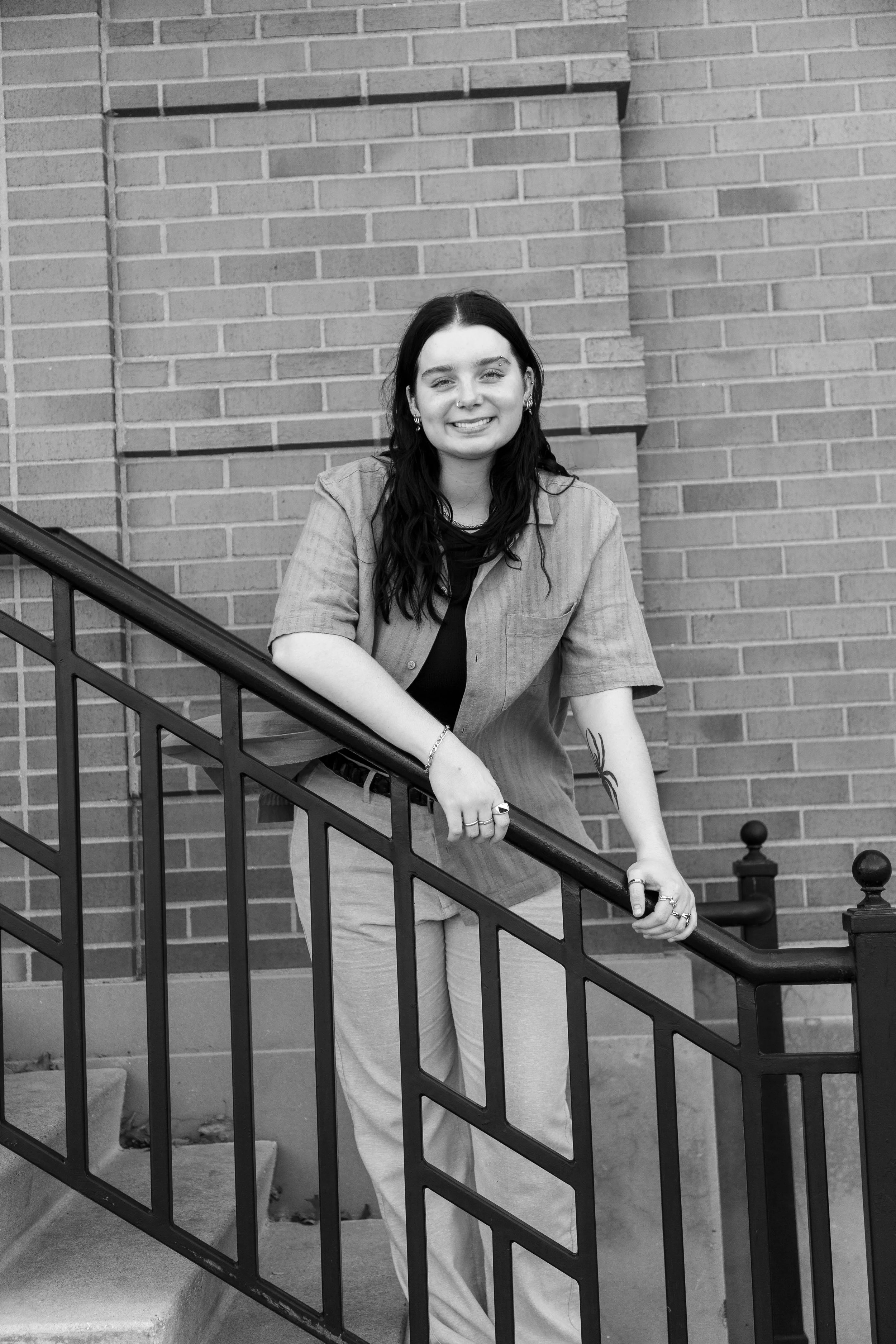 Young woman with dark hair smiling, wearing a short-sleeved shirt and light-colored pants, standing on stairs holding a railing against a brick wall.