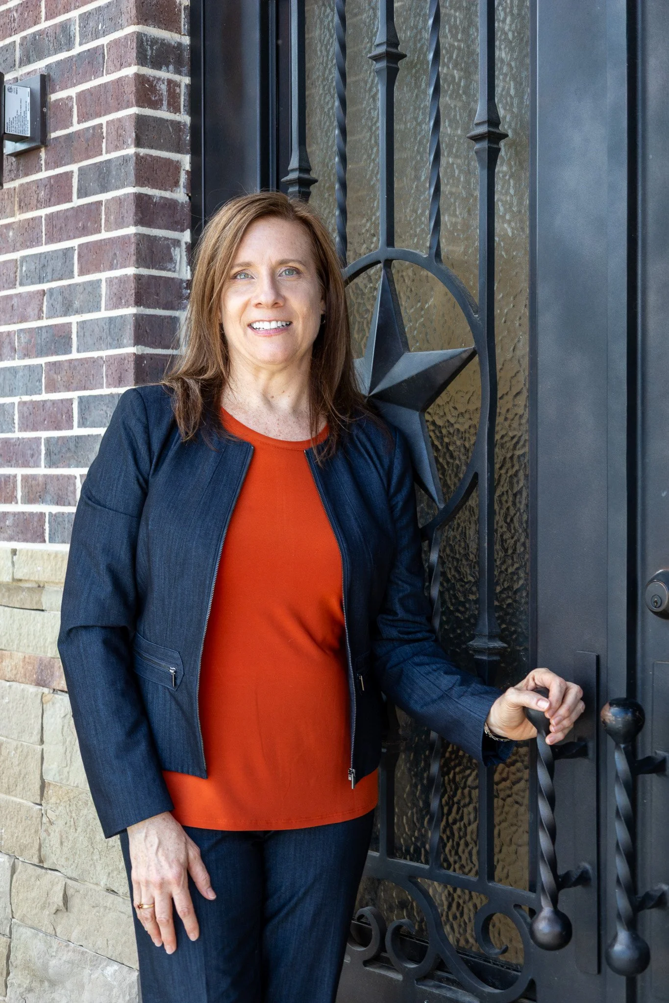 A woman with brown hair and fair skin standing outdoors near a brick building, holding the door handle of a black decorative door with a star on it.