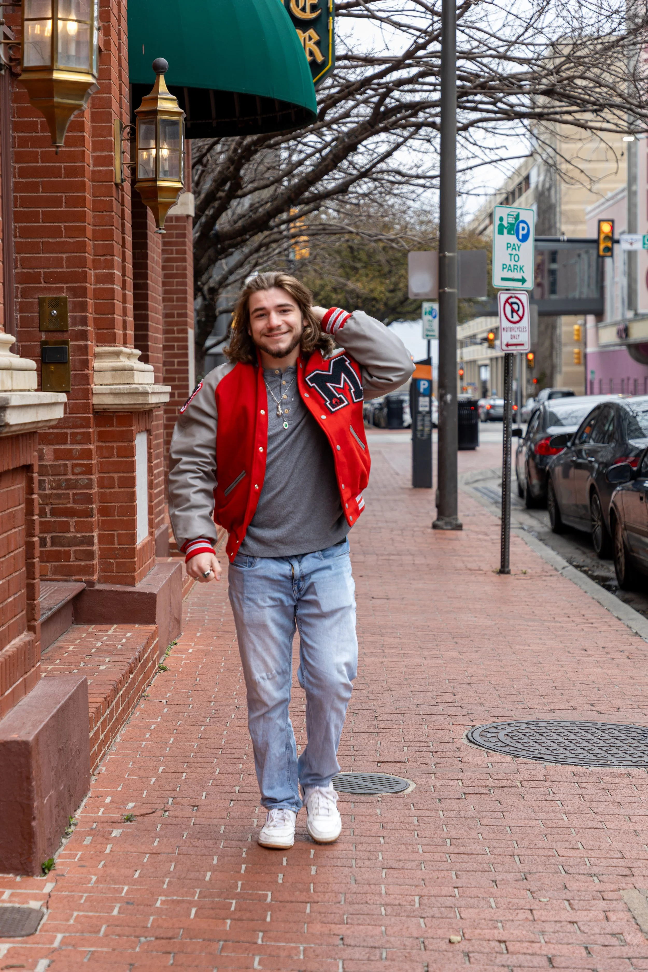 A young man walking down a brick sidewalk in an urban setting, smiling, wearing a red and gray varsity jacket, gray shirt, light blue jeans, and white sneakers, with trees and parked cars in the background.