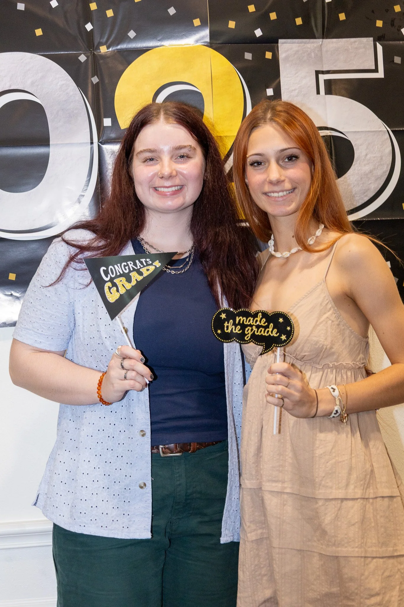 Two women at a graduation celebration holding signs that say "Congrats Grad" and "Made the Grade". They are smiling and standing in front of a black and gold backdrop.