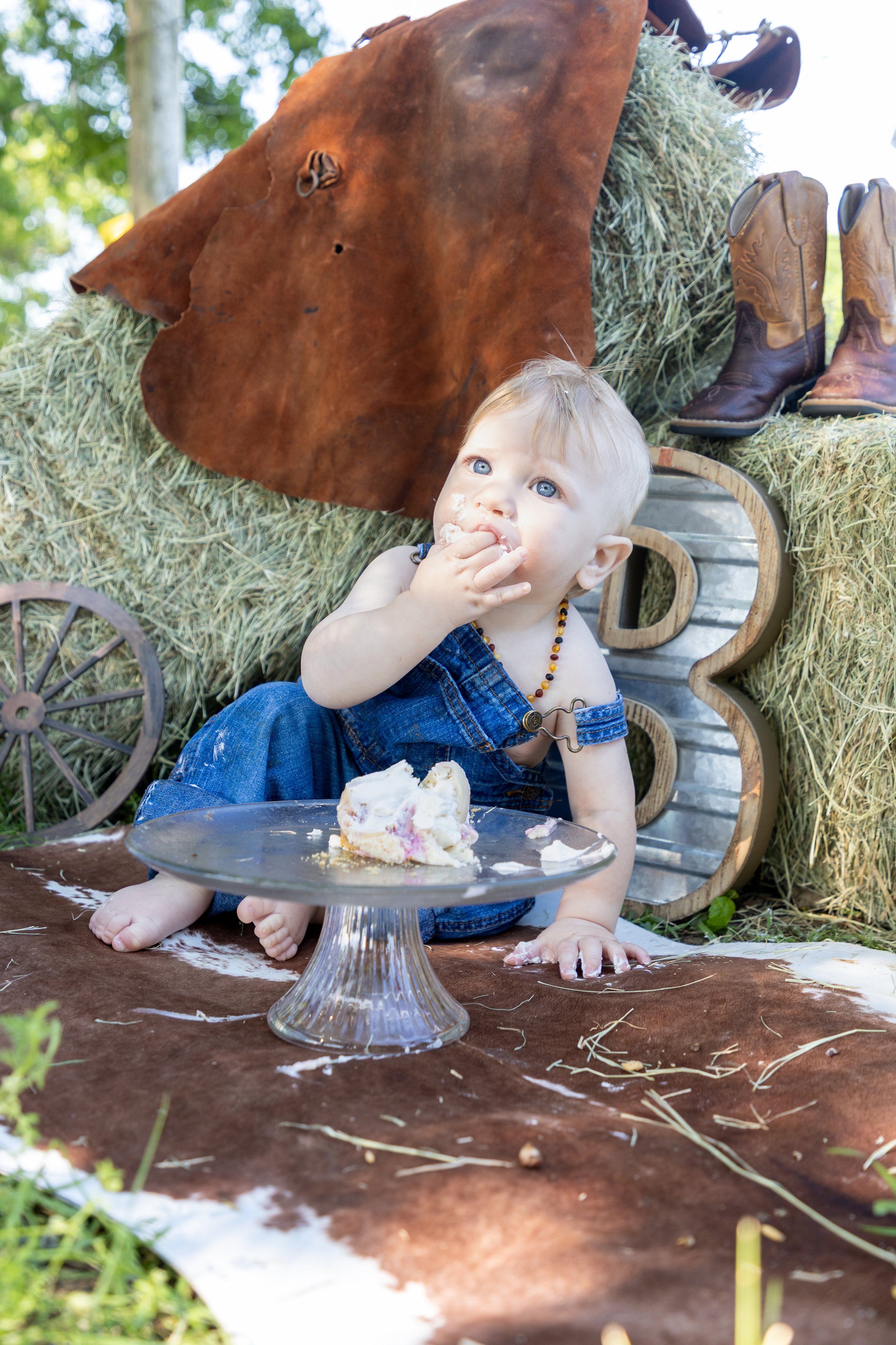 A young child sitting on a animal hide, eating cake from a cake stand during a farm-themed party. There are cowboy boots on hay bales behind him and a large animal leather piece hanging nearby.