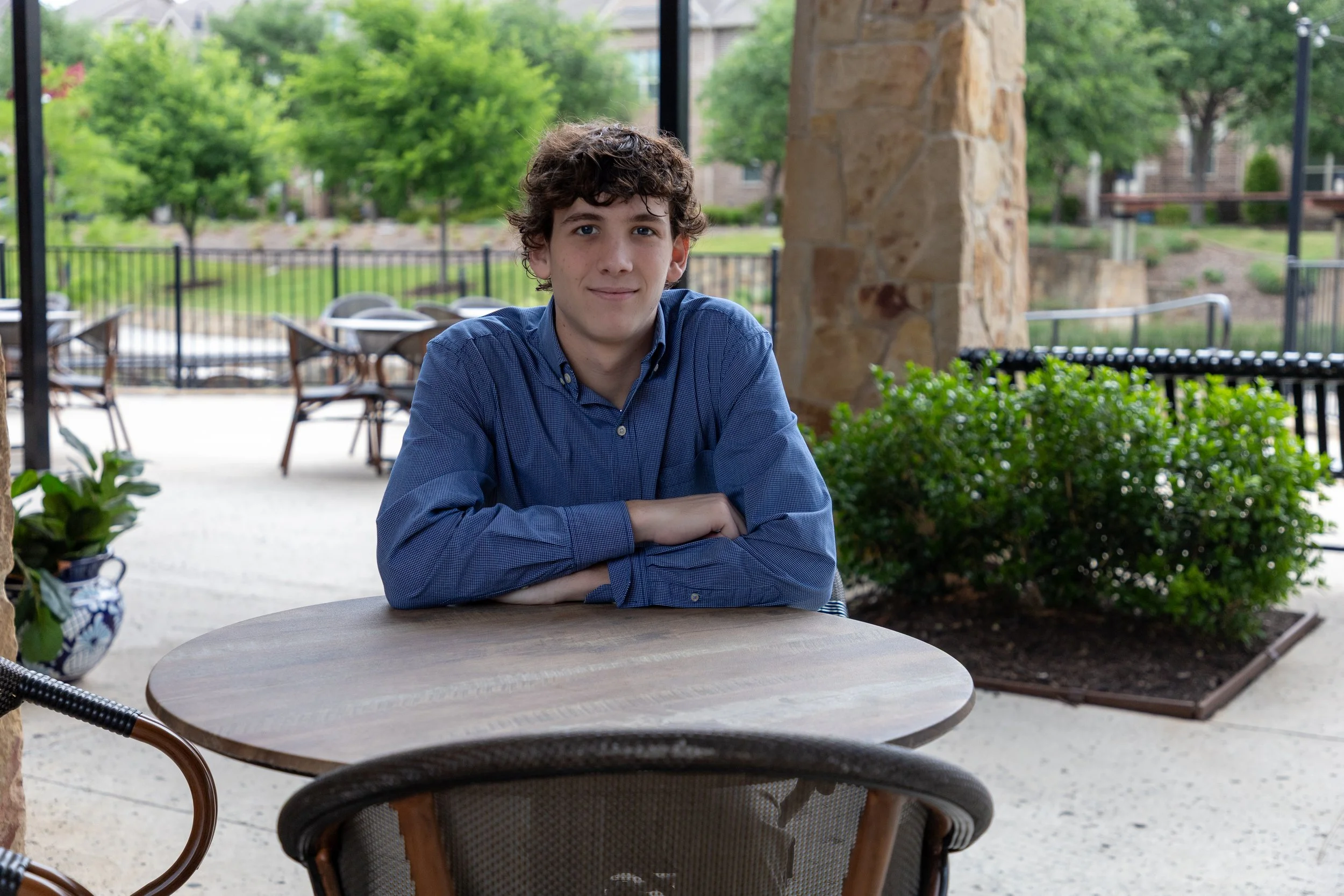 A young man with curly brown hair and a blue button-up shirt sitting at an outdoor cafe table with greenery and trees in the background.