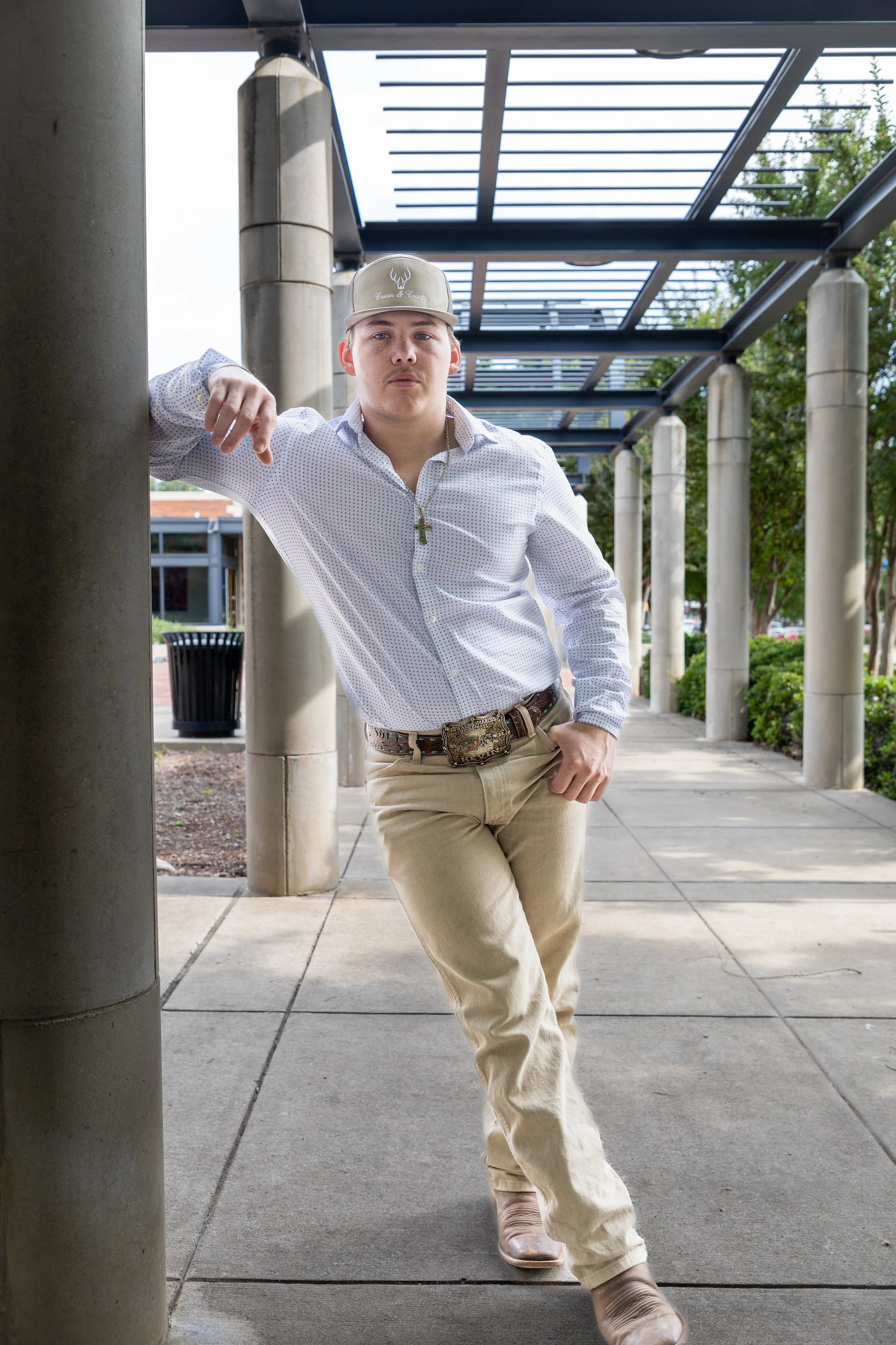 A young man wearing a light-colored baseball cap, white patterned shirt, beige pants, and cowboy boots standing outdoors under a covered walkway, leaning against a column with one hand in his pocket.