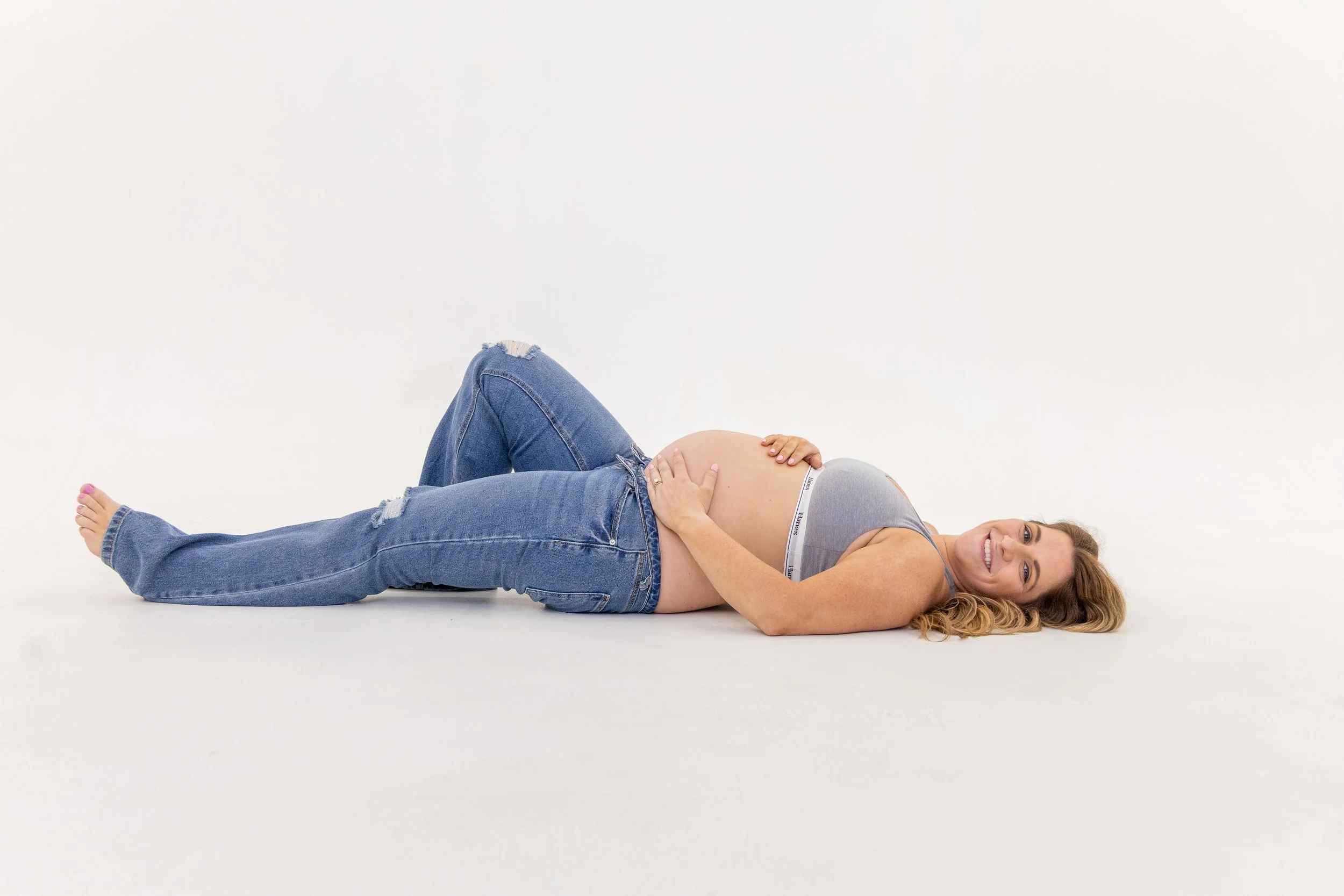 A pregnant woman lying on a white floor, smiling, with her head turned to the side and her hand resting on her belly, wearing ripped jeans and a grey tank top.