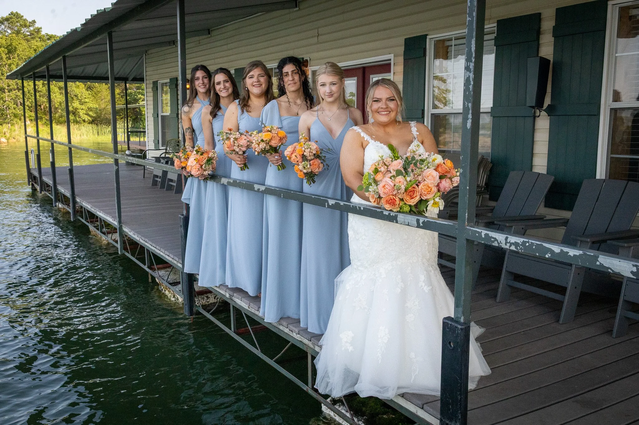 A bride and five bridesmaids standing on a dock over a body of water, holding bouquets of pink, peach, and orange flowers. The bride is in a white wedding gown, and the bridesmaids are in matching light blue dresses. They are smiling for the photo.