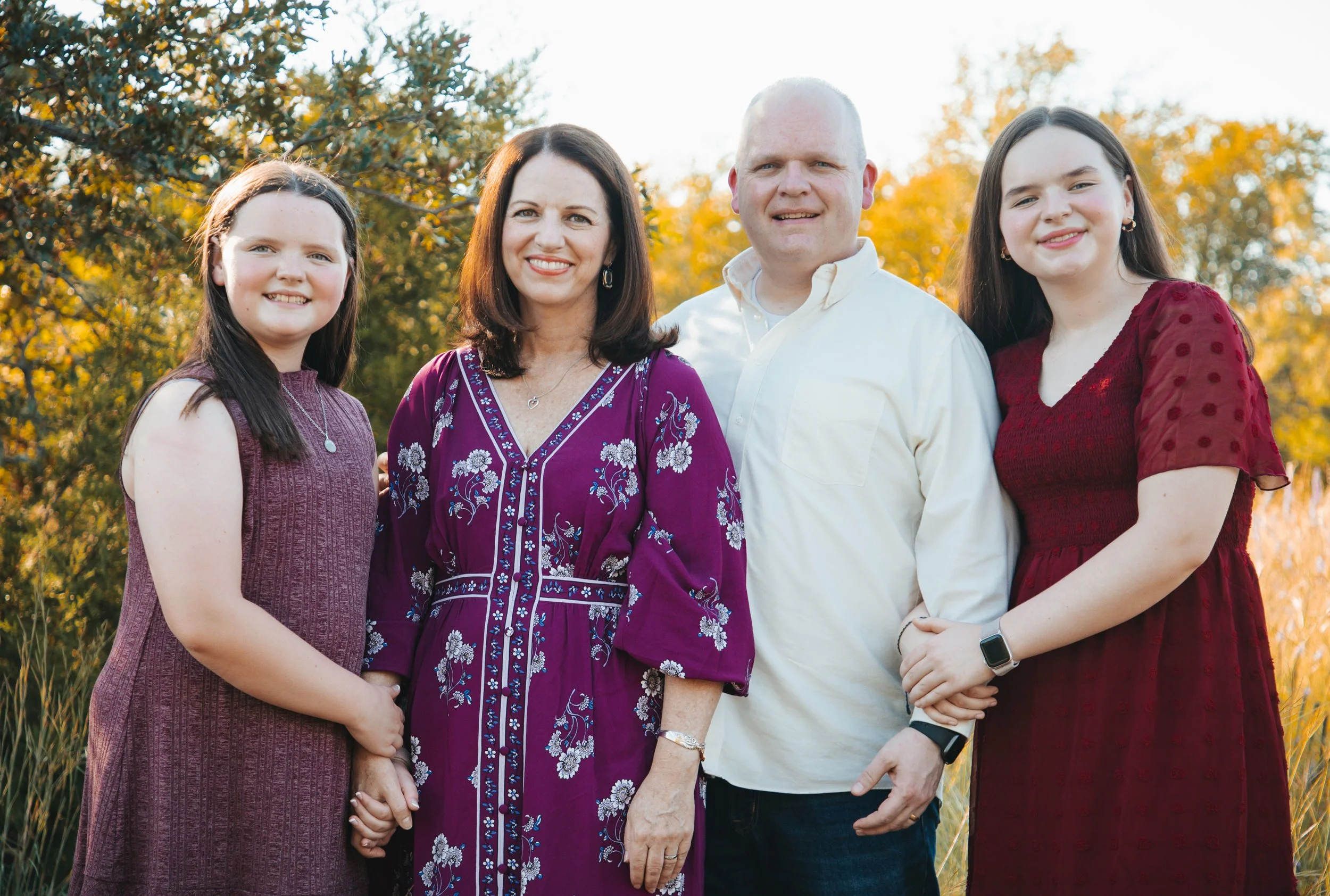 Family of five outdoors during autumn, standing in front of trees with fall foliage. The family includes two young women, a woman, a man, and a girl, all smiling at the camera.