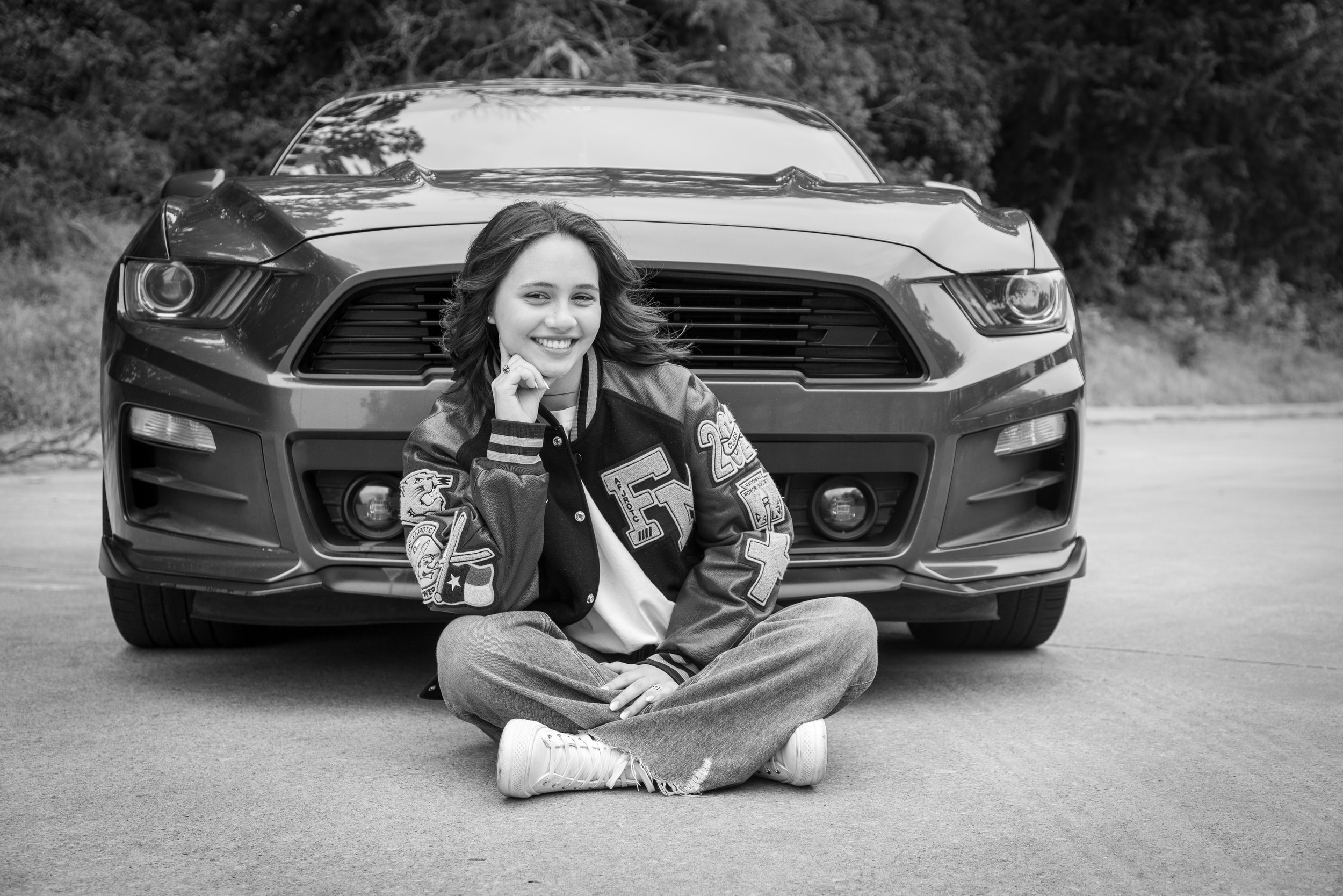 A girl sitting cross-legged on the ground in front of a large car, smiling and looking at the camera.