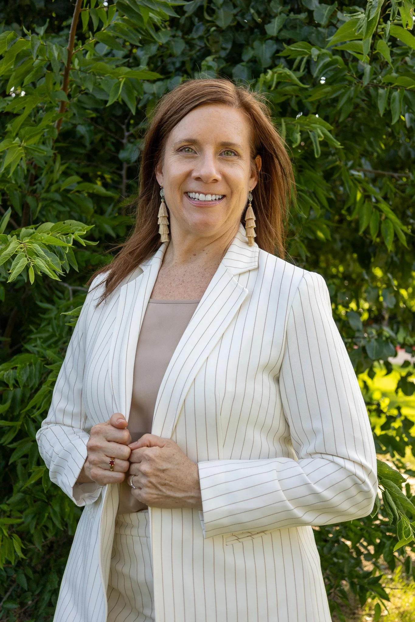 A woman with long reddish brown hair, wearing a white pinstripe blazer and beige top, standing outdoors in front of green foliage, smiling at the camera.