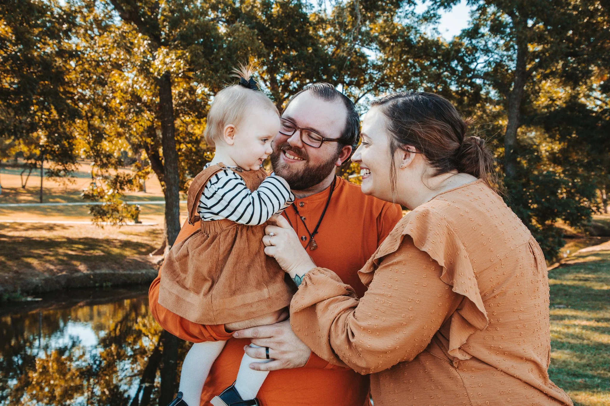 A family outdoors during autumn, with a young girl holding her parents' faces and smiling, surrounded by trees with fall foliage.
