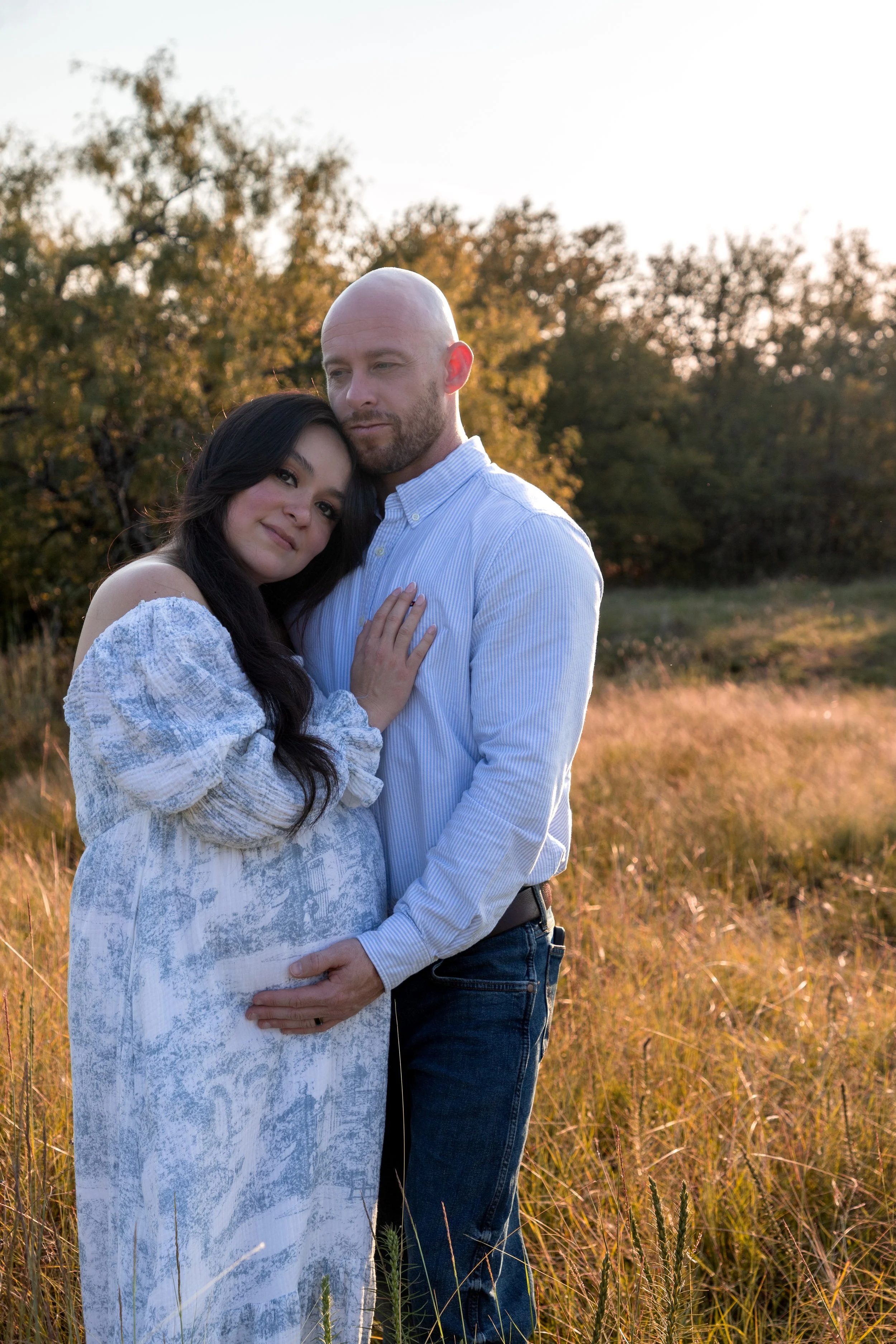 A woman and a man embrace in a field at sunset, with trees in the background.