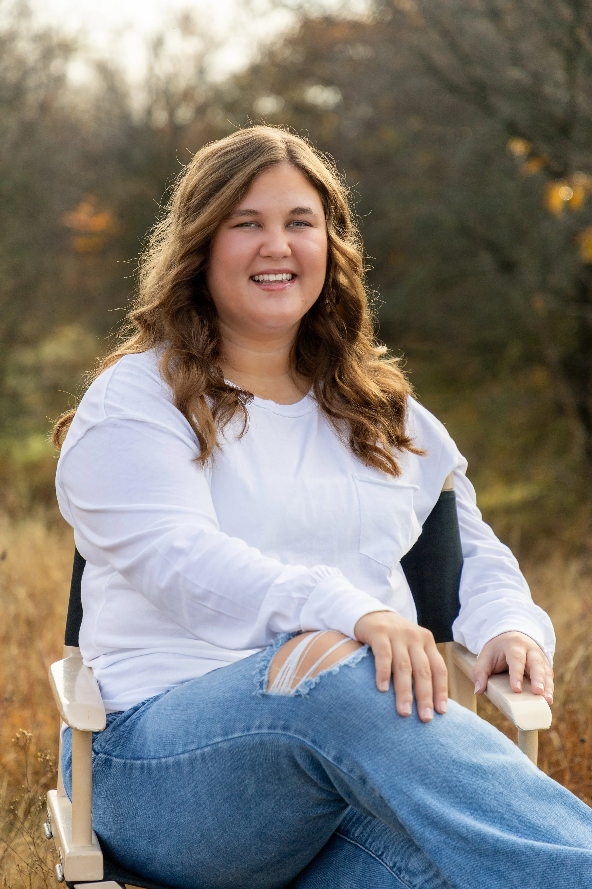 A smiling woman with long wavy brown hair sitting outdoors in a park during fall, wearing a white long sleeve shirt and ripped blue jeans.