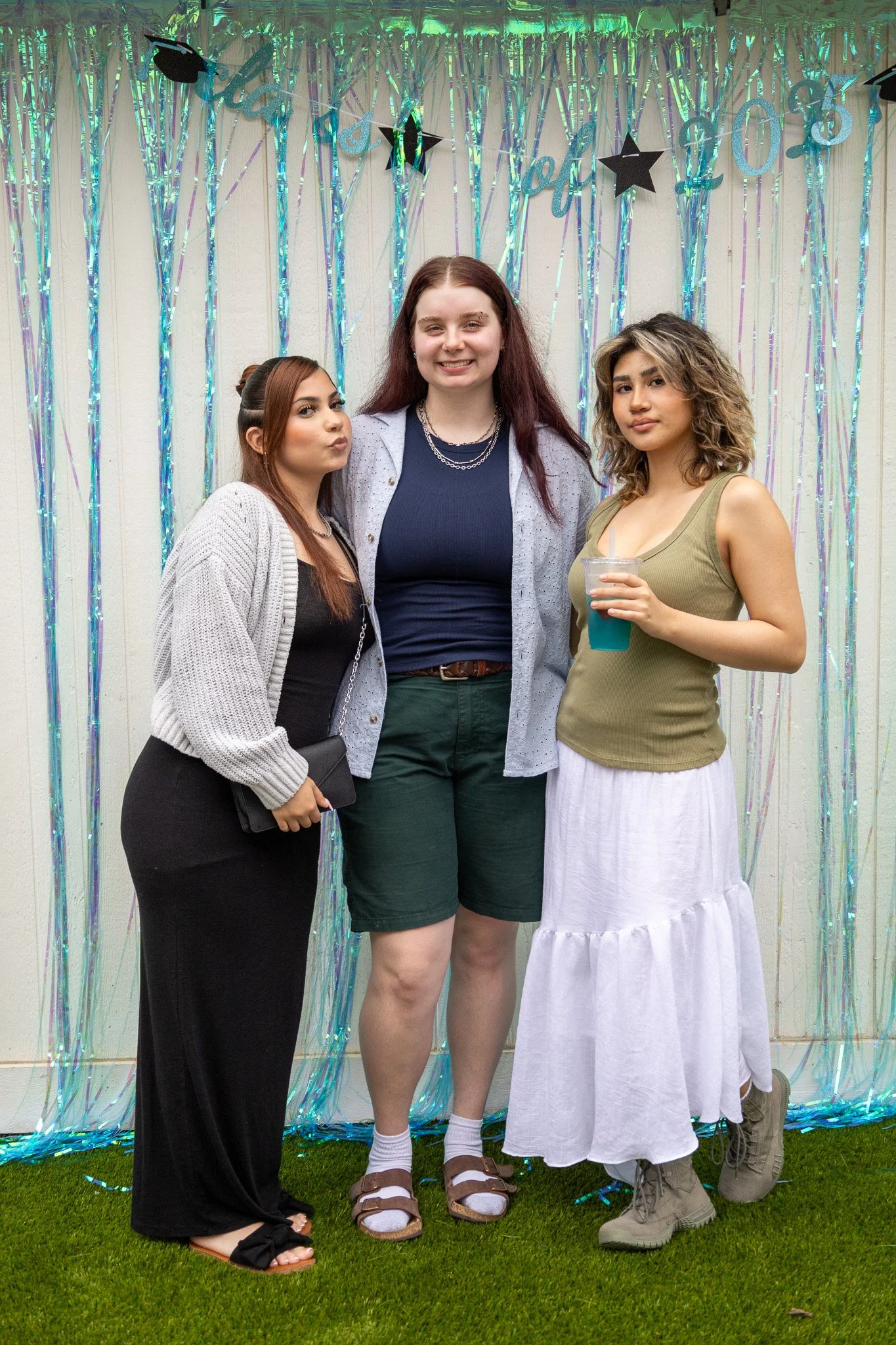 Three women standing together at a celebration, with shiny teal and purple streamers and black star decorations in the background, on a grass surface.