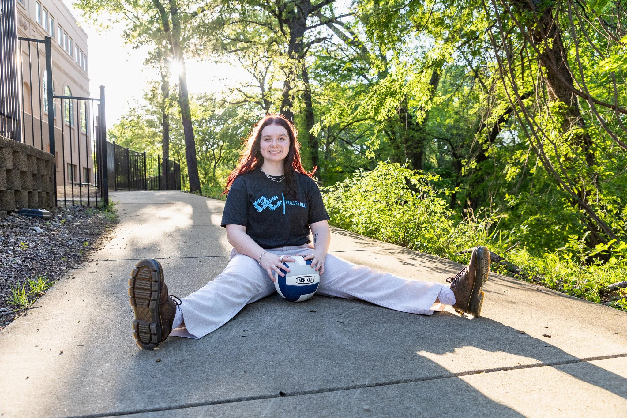 A young woman with red hair and a black T-shirt sitting on the sidewalk with her legs spread apart and holding a volleyball, smiling at the camera, surrounded by trees and sunlight.