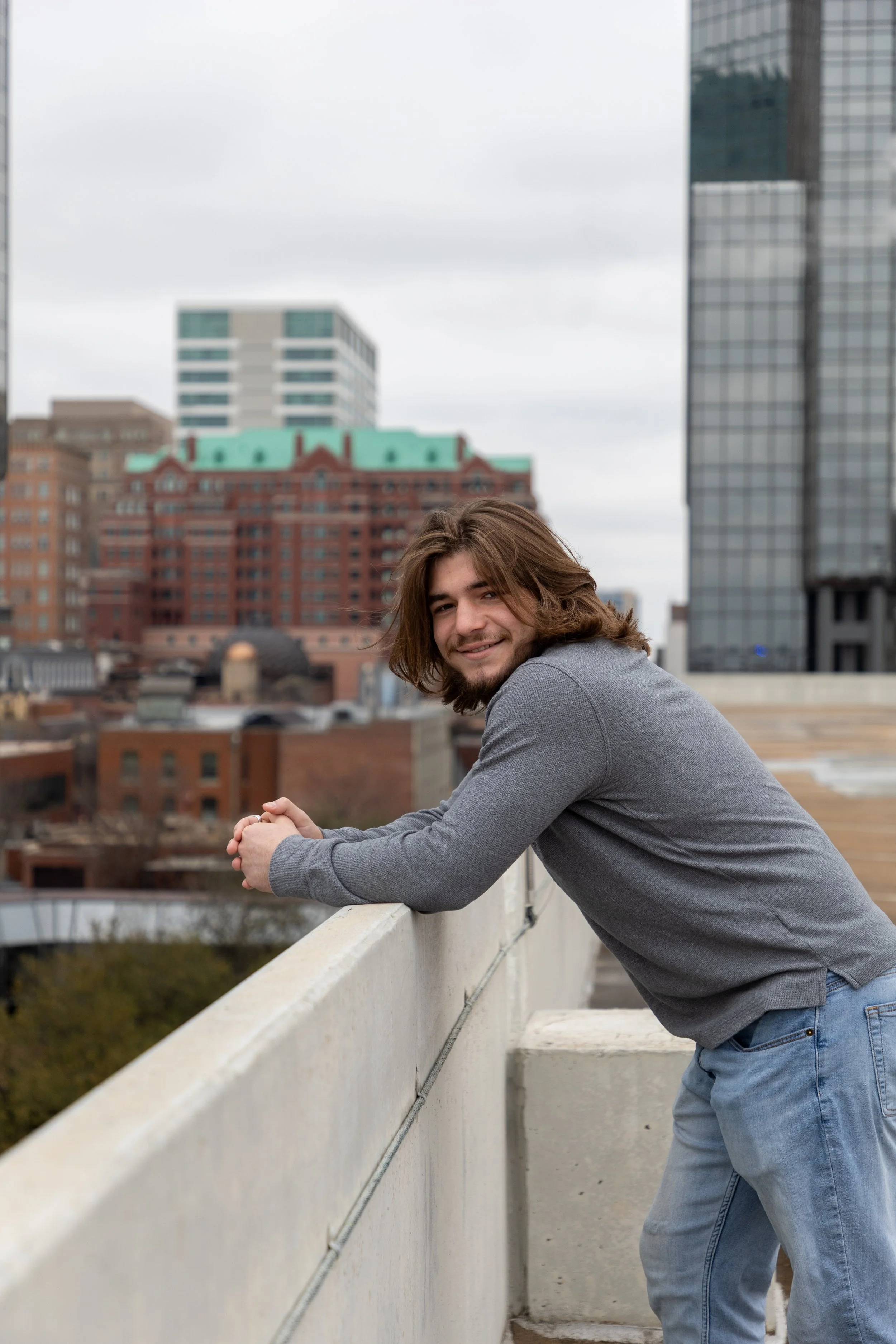 Young man with shoulder-length brown hair and beard leaning on a rooftop railing, smiling, with a cityscape of tall buildings in the background under gray sky.