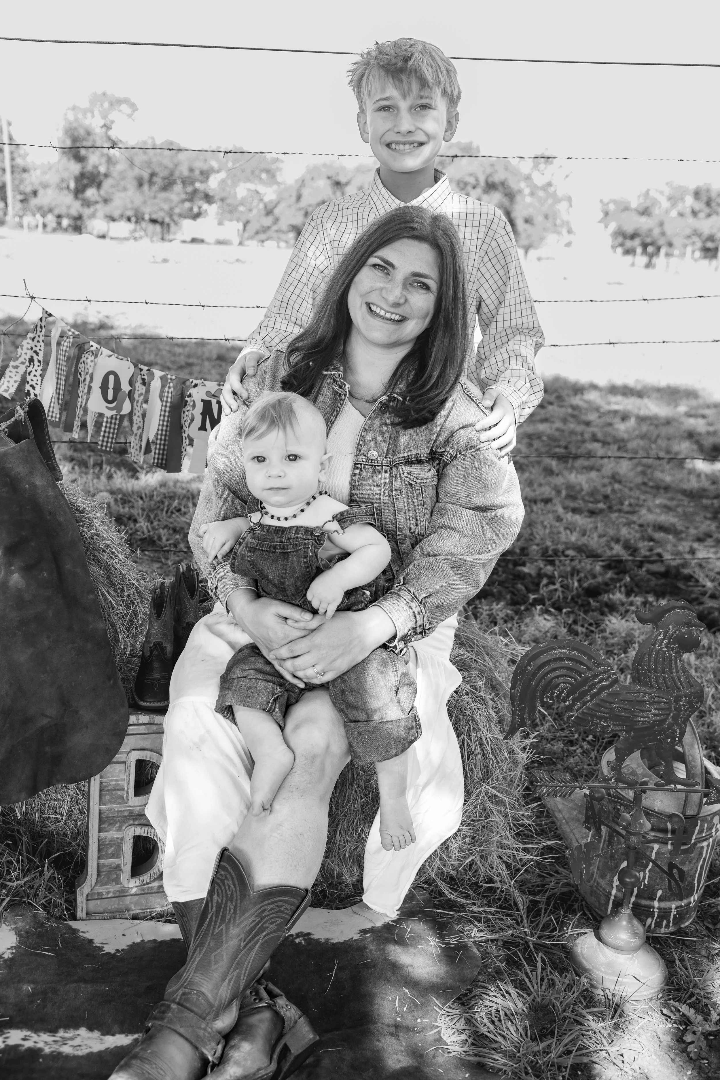 A woman with two children sitting outdoors with a barnyard theme, barbed wire in the background, and decorative rooster and banner, in black and white.