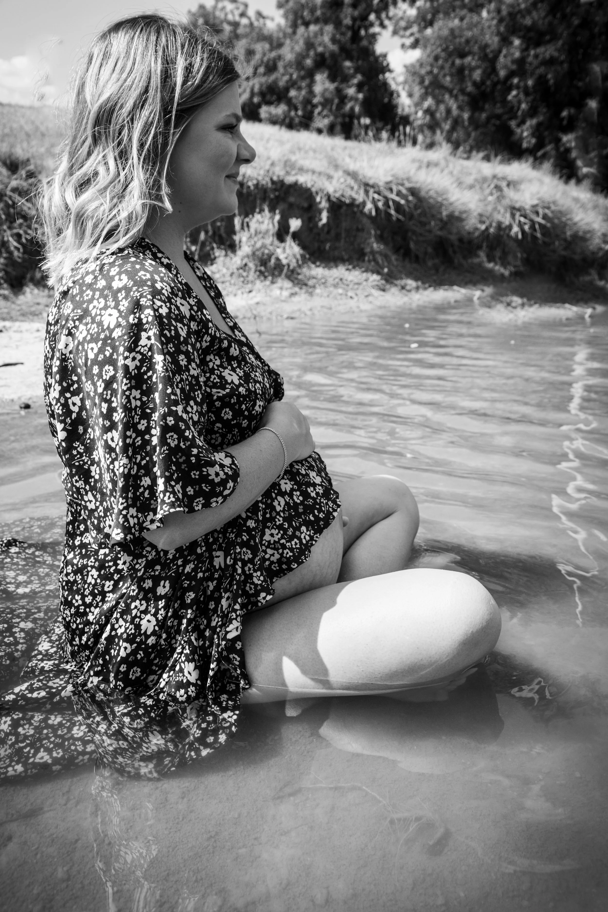 A pregnant woman in a floral dress sitting on a rock with her feet in the water at a beach or riverside, smiling and holding her belly.