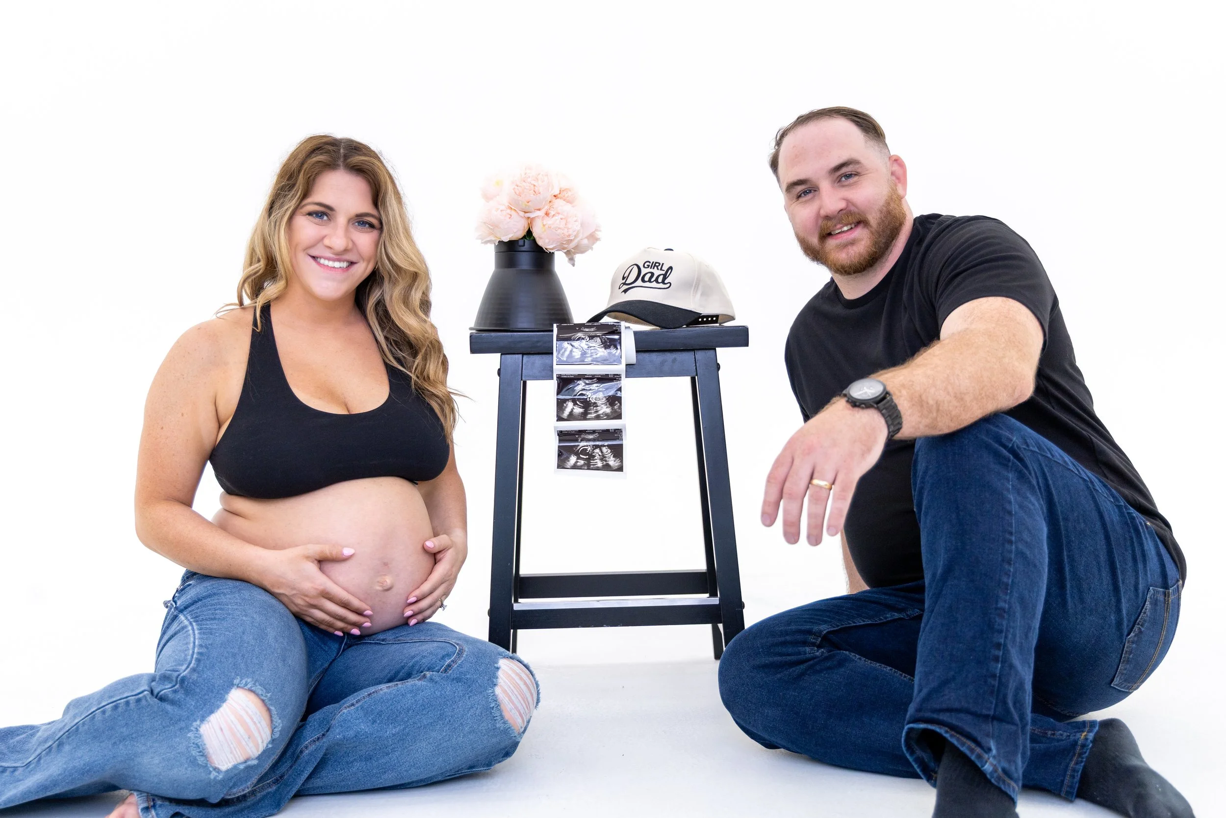A pregnant woman with long wavy hair smiling, sitting on the floor with her legs crossed, wearing a black tank top and ripped jeans, holding her belly. A man with a beard and short hair smiling, kneeling on one knee, wearing a black T-shirt and jeans