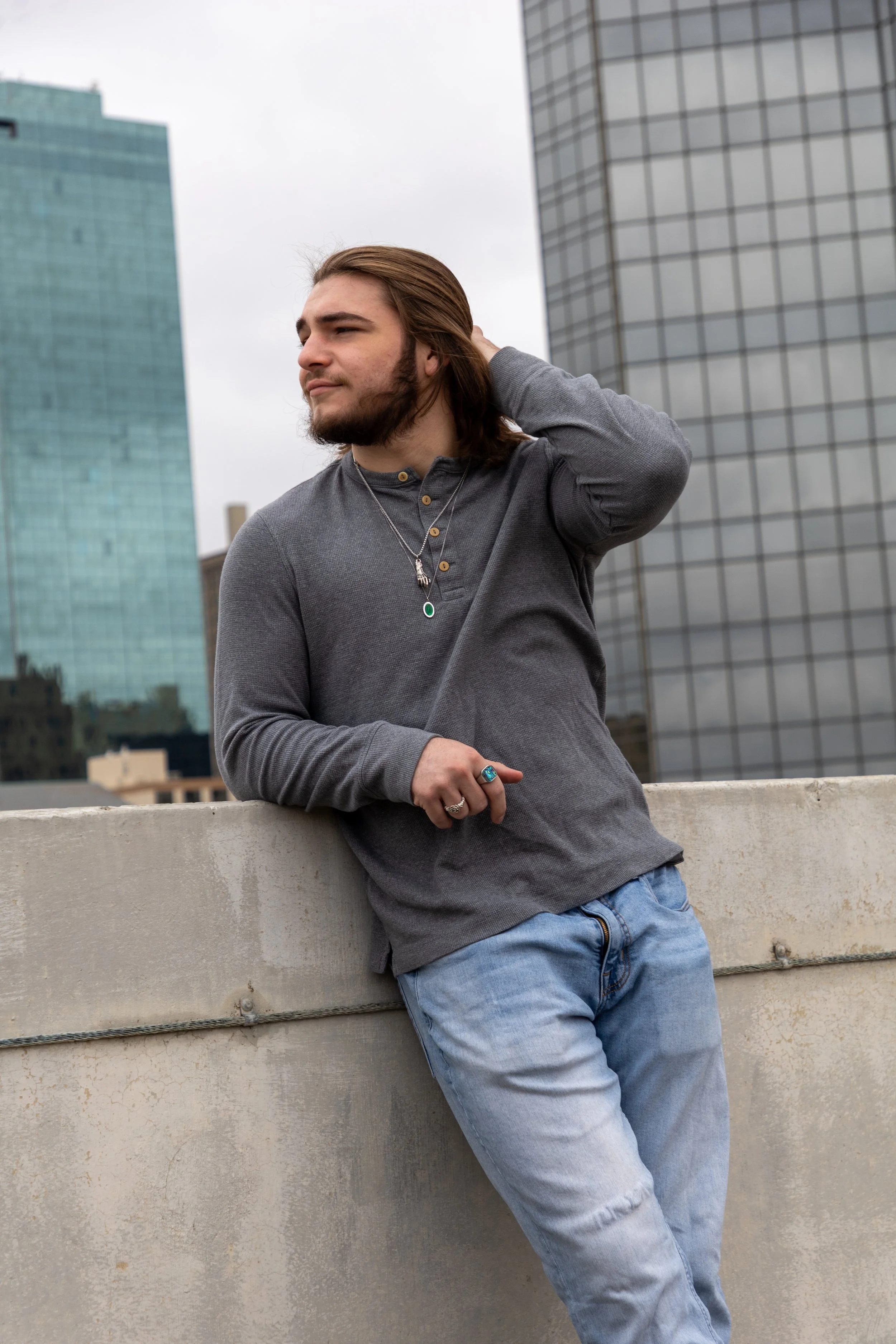A young man with long brown hair and a beard, wearing a gray long-sleeve shirt and light blue jeans, leaning on a concrete ledge on a rooftop with tall modern glass buildings in the background.