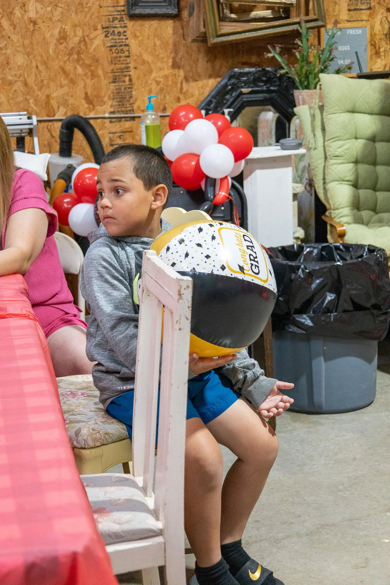 A young boy sitting on a floral cushioned chair holding a beach ball, at a birthday party with balloons and festive decorations in the background.