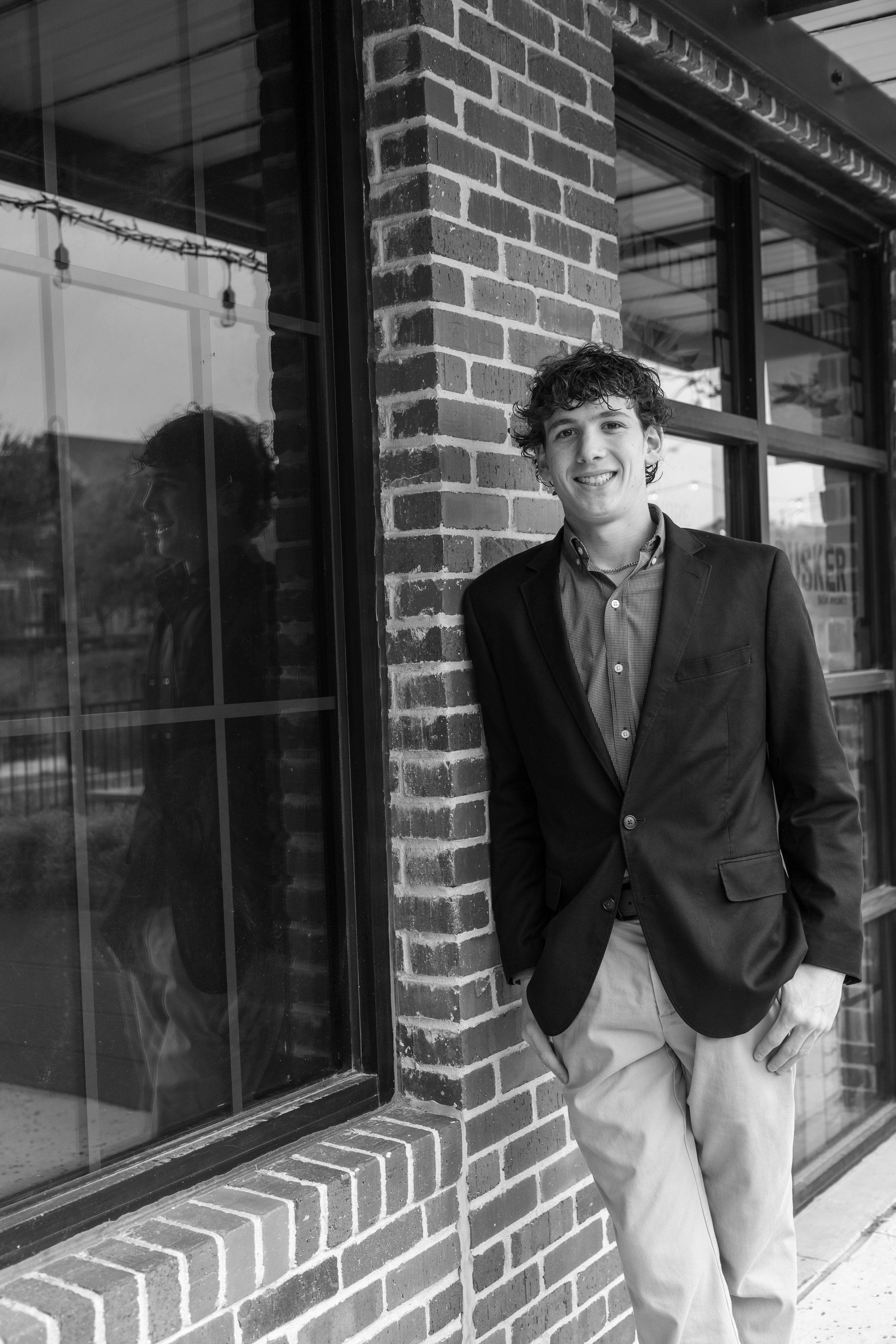 A young man with curly hair wearing a blazer and light-colored pants, standing outside next to a glass window of a brick building, smiling at the camera, with his reflection visible in the window.
