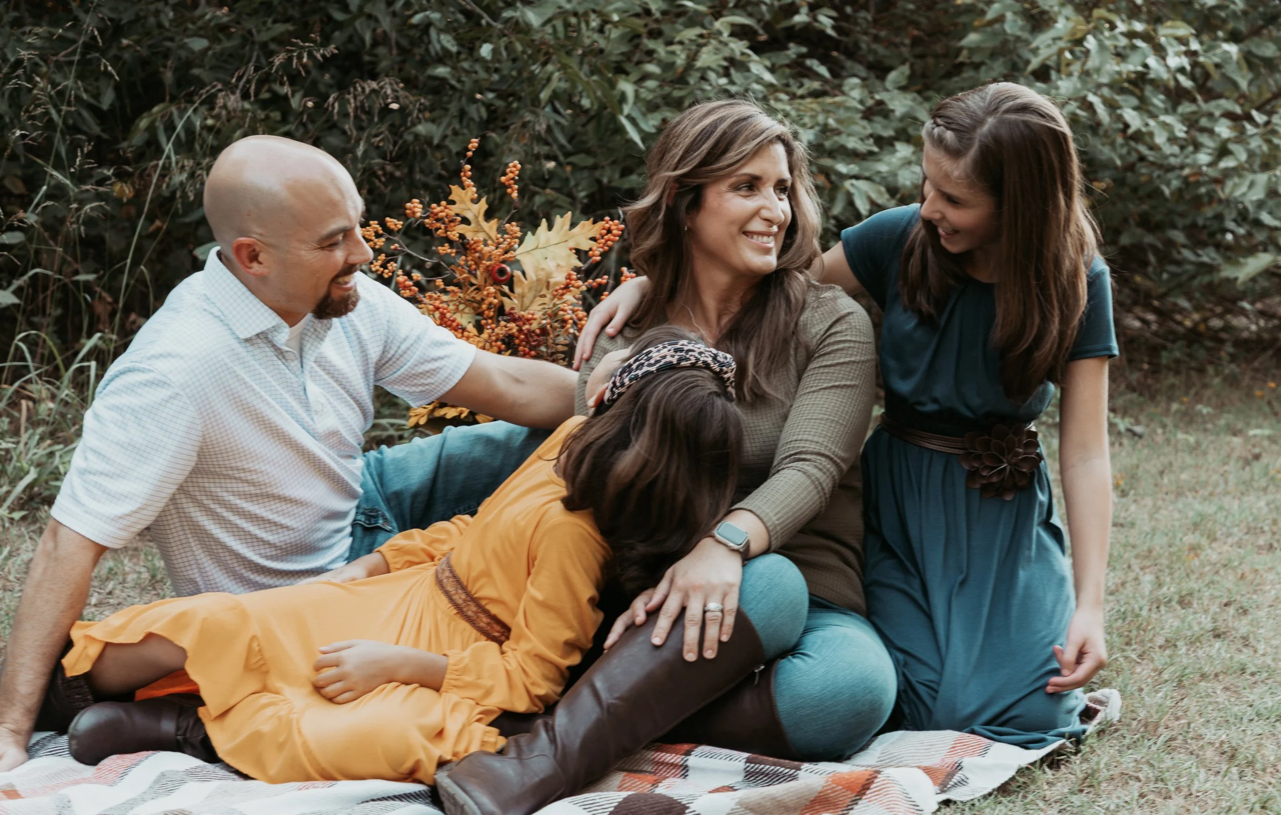 Family enjoying fall outdoors, a woman sitting on blanket with two kids and a man, all smiling and affectionate.