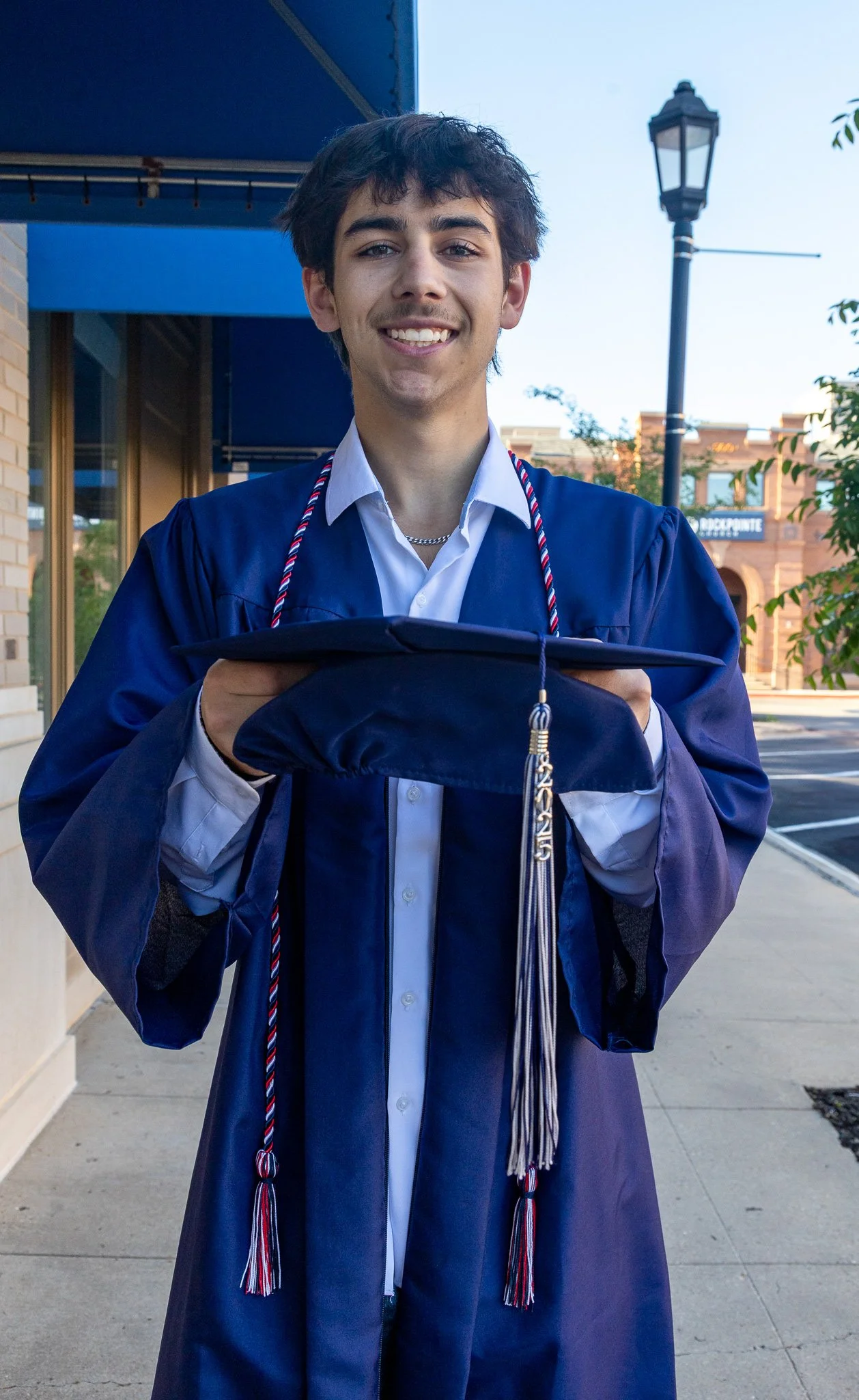 A young man in a graduation gown holding a cap and smiling outdoors.