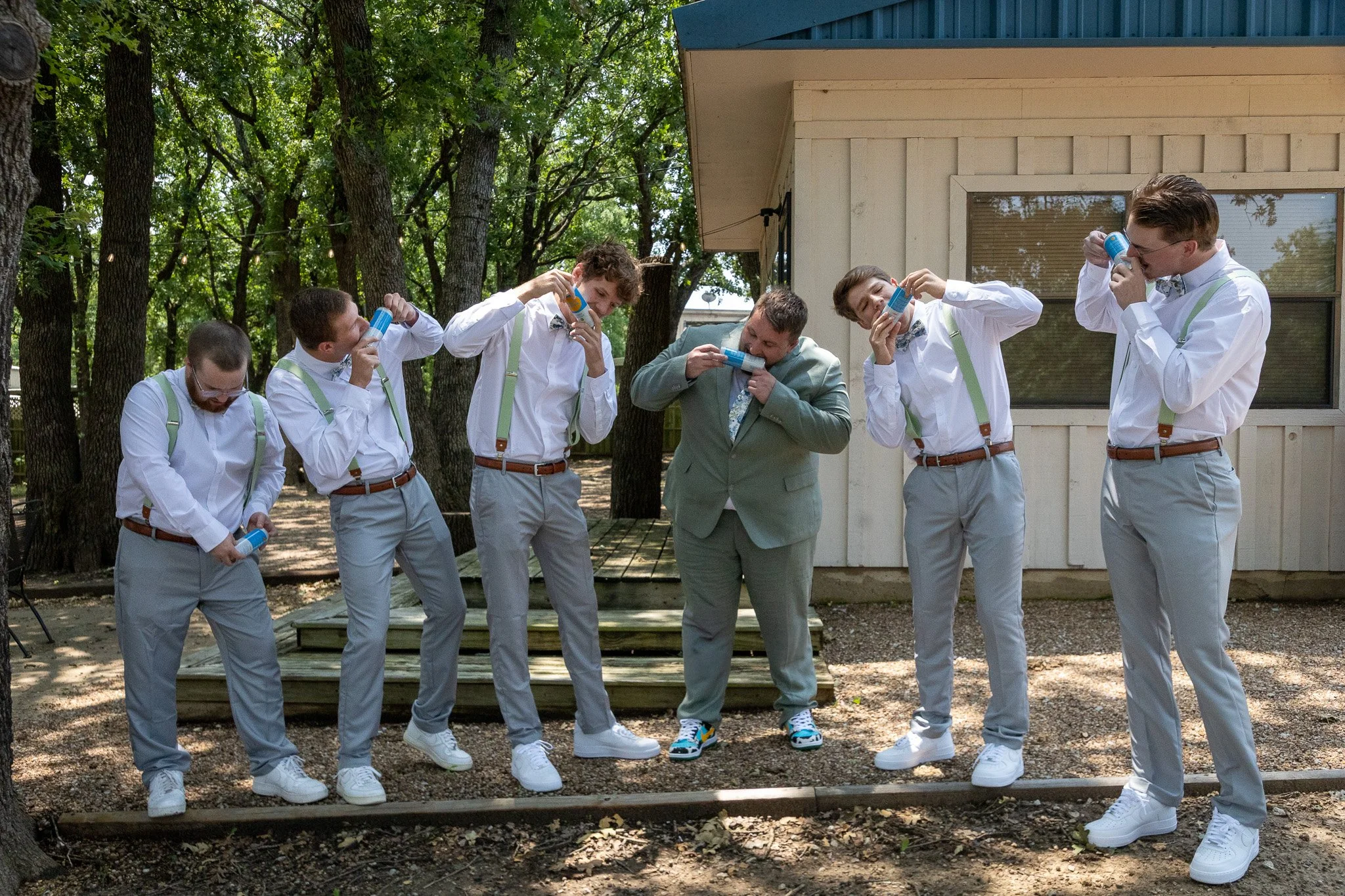 Group of six men dressed in white shirts and grey pants, standing outdoors near a white building and trees, using multiple small breathalyzer devices.