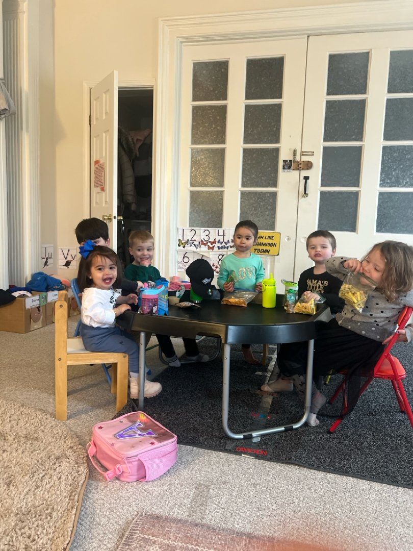Children sitting around a table in a classroom setting, engaged in activities. There's a pink backpack on the floor and educational posters on the wall.