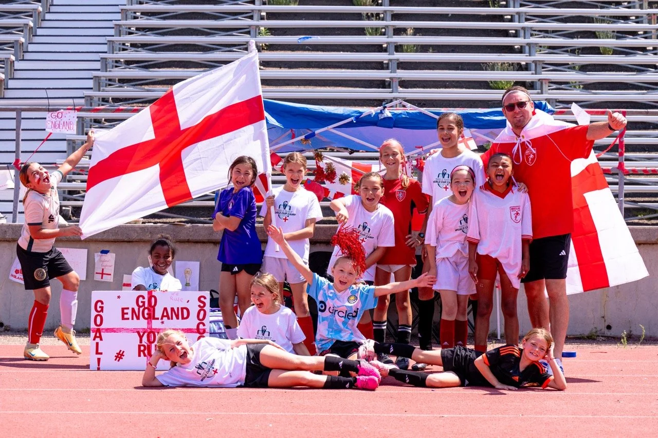 A group of young girls and their coach celebrating on a sports field, holding flags and wearing England national soccer team jerseys, with cheering expressions and posing for a photo.