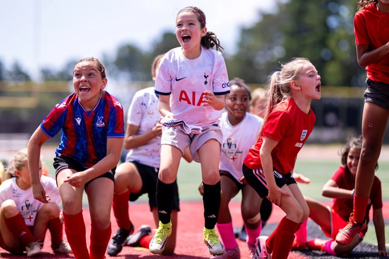 Group of young girls playing and laughing on a sports field, some lifting themselves off the ground in a playful activity.