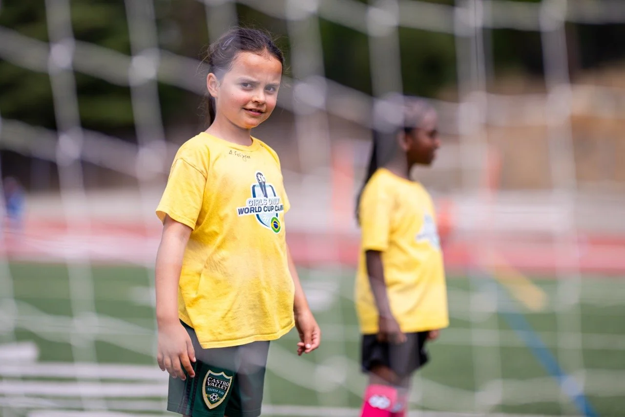 Two young girls in yellow soccer shirts standing on a soccer field, with one girl in the foreground and the other girl in the background. They are behind a goal net, and one girl has dark hair tied back, while the other has braids.