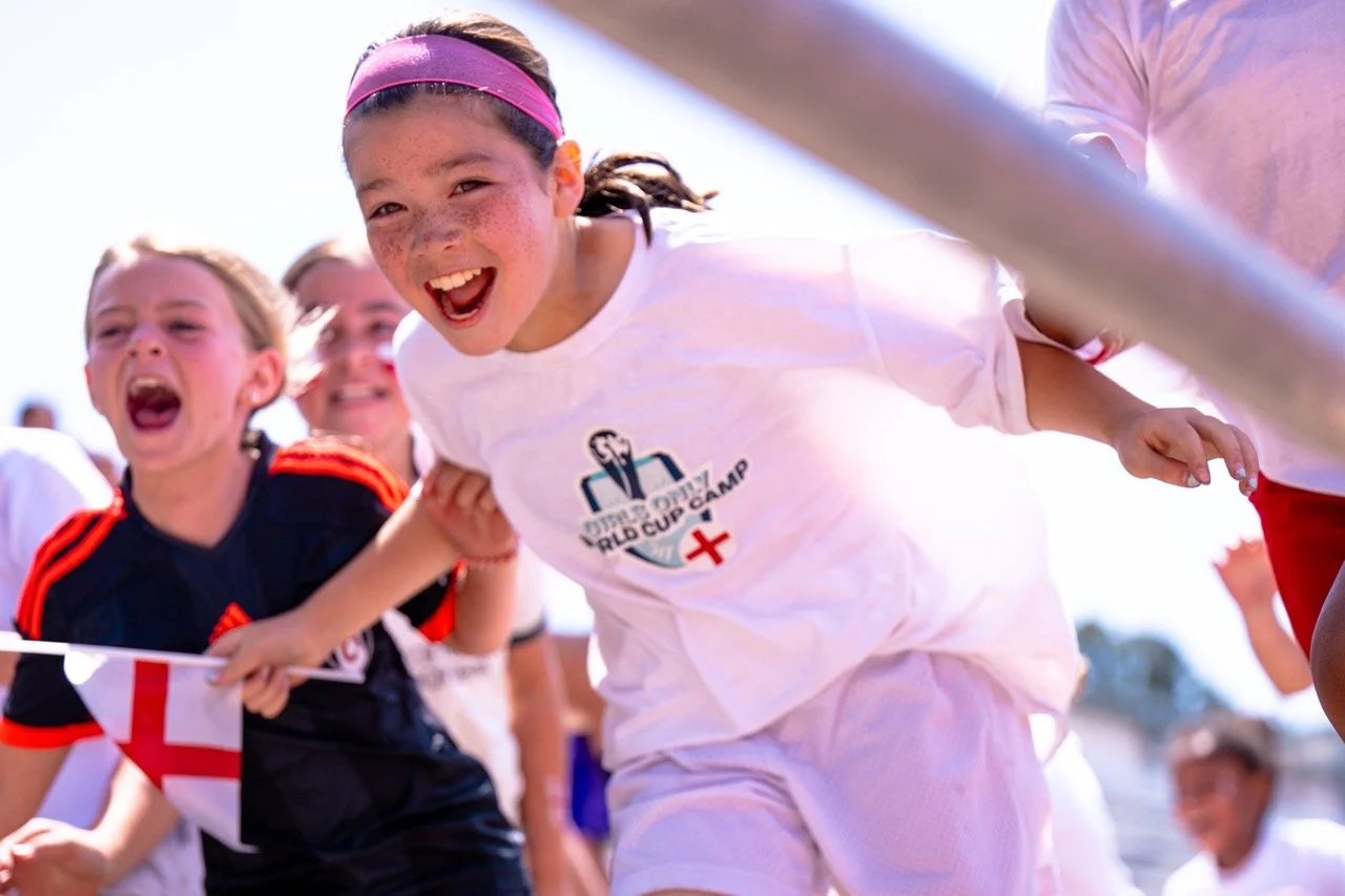 Children participating in a race at a sports camp, holding hands and smiling, with a girl wearing a white t-shirt and pink headband leading.