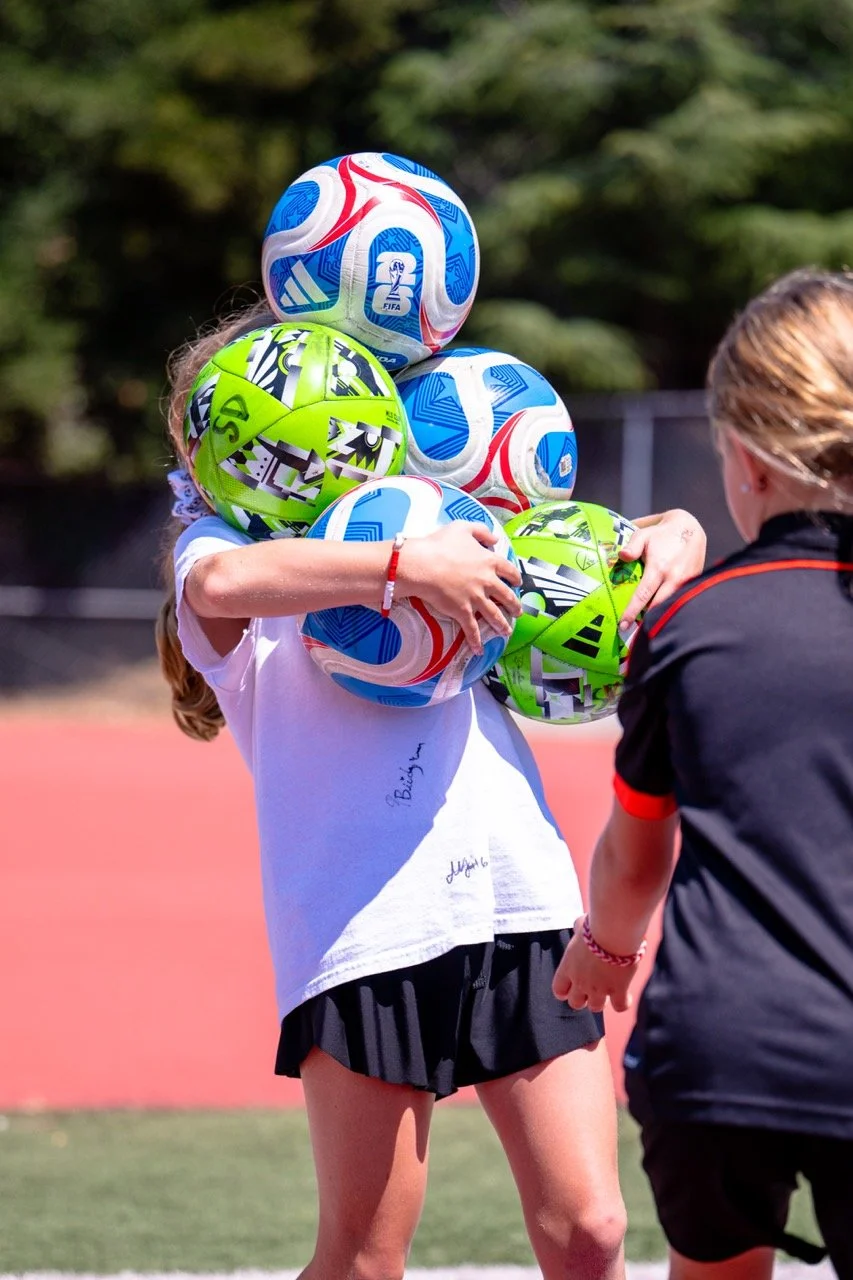 Child holding multiple colorful soccer balls on an outdoor sports field with another person nearby.