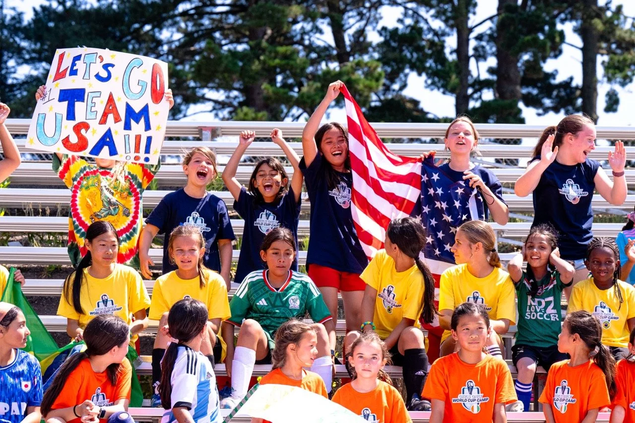 Group of young girls sitting and standing on bleachers at a sports event, some wearing yellow t-shirts, others in soccer jerseys, with flags and a sign that says 'Let's Go USA' in the background.