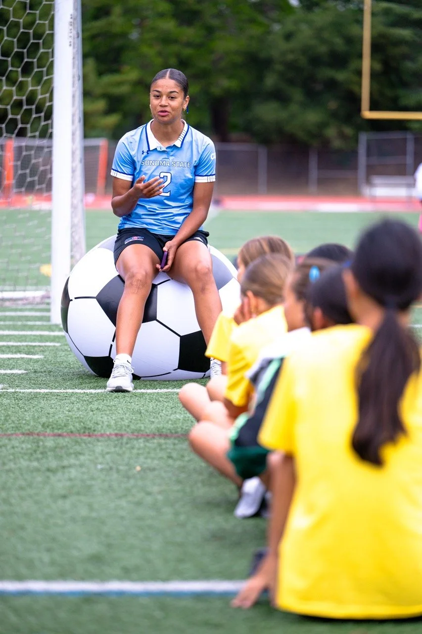 A young female soccer player in a blue jersey with the number 2, sitting on an inflatable soccer ball on a soccer field, speaking to a group of children sitting on the ground in yellow jerseys.