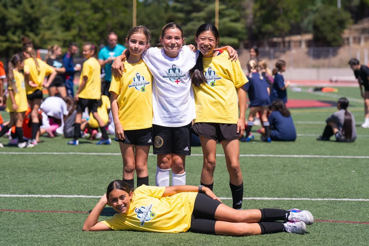Four young girls at a sports field, three standing with their arms around each other and one lying on the ground in front, all smiling. They are wearing sports uniforms, likely for soccer or rugby, during a youth sports event.