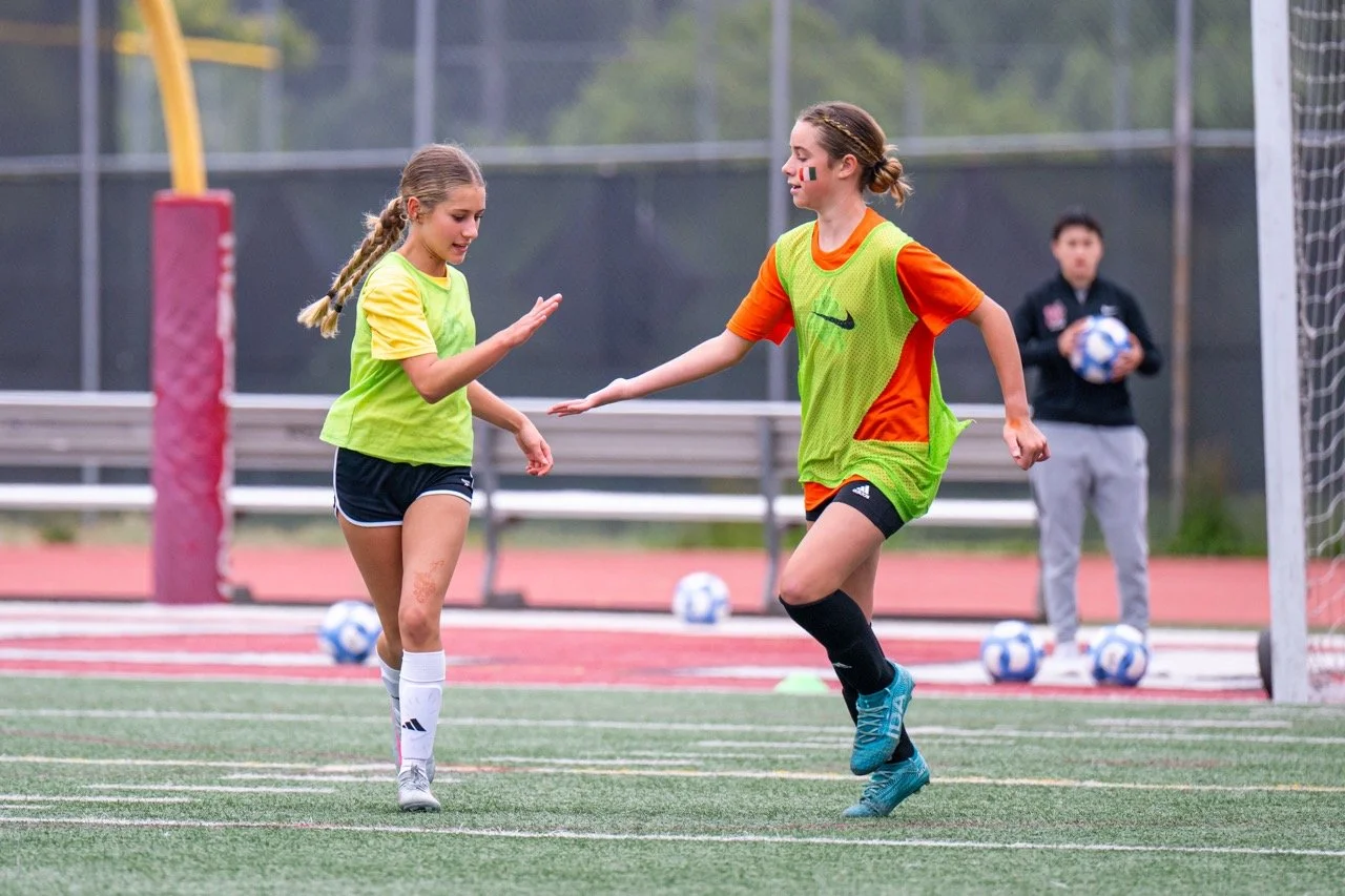 Two young female soccer players in training, one giving a high five, on a soccer field with a coach holding a soccer ball in the background.