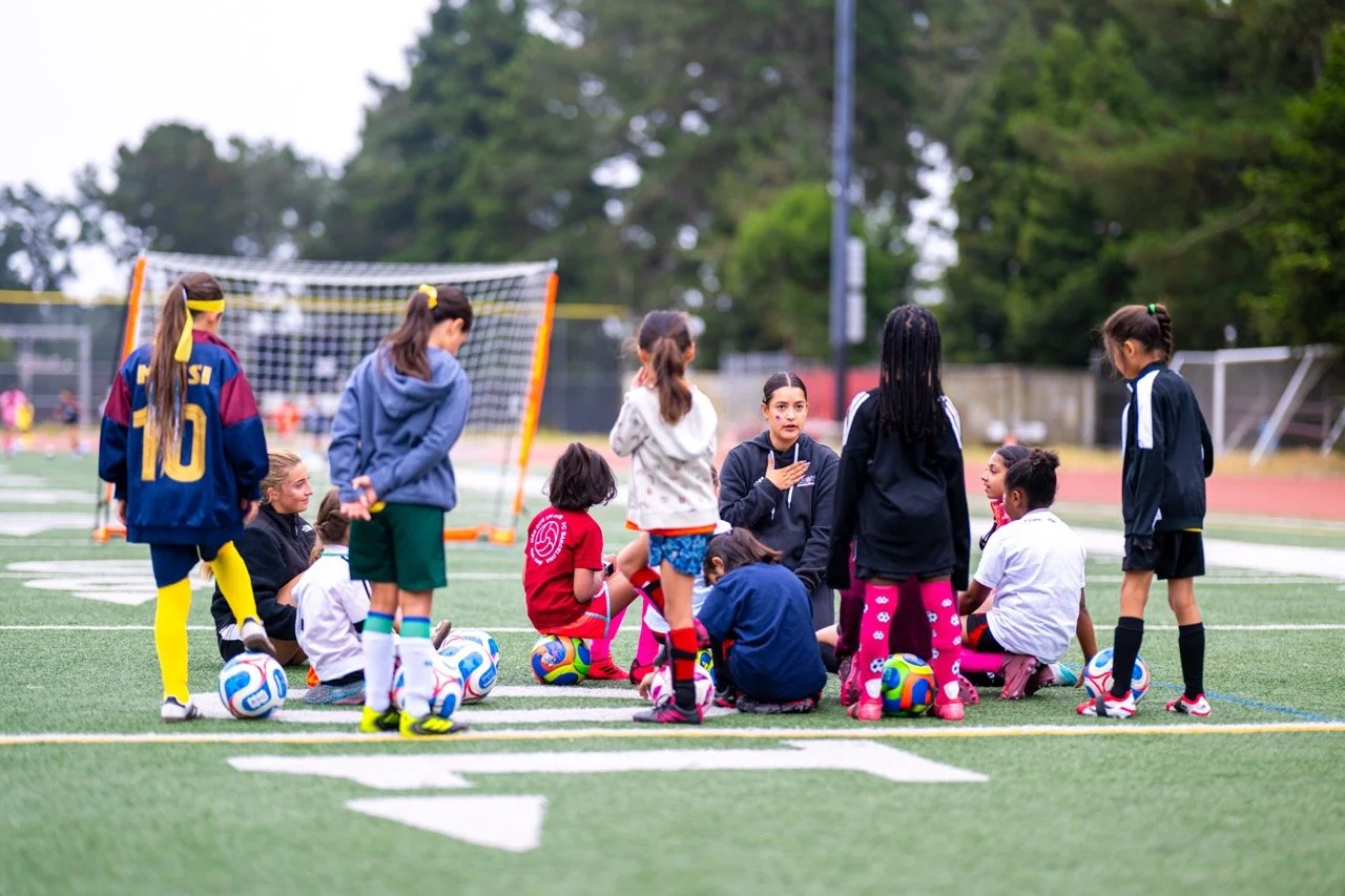 Group of young girls on a soccer field, listening to a coach during a training session, with goalposts and trees in the background.