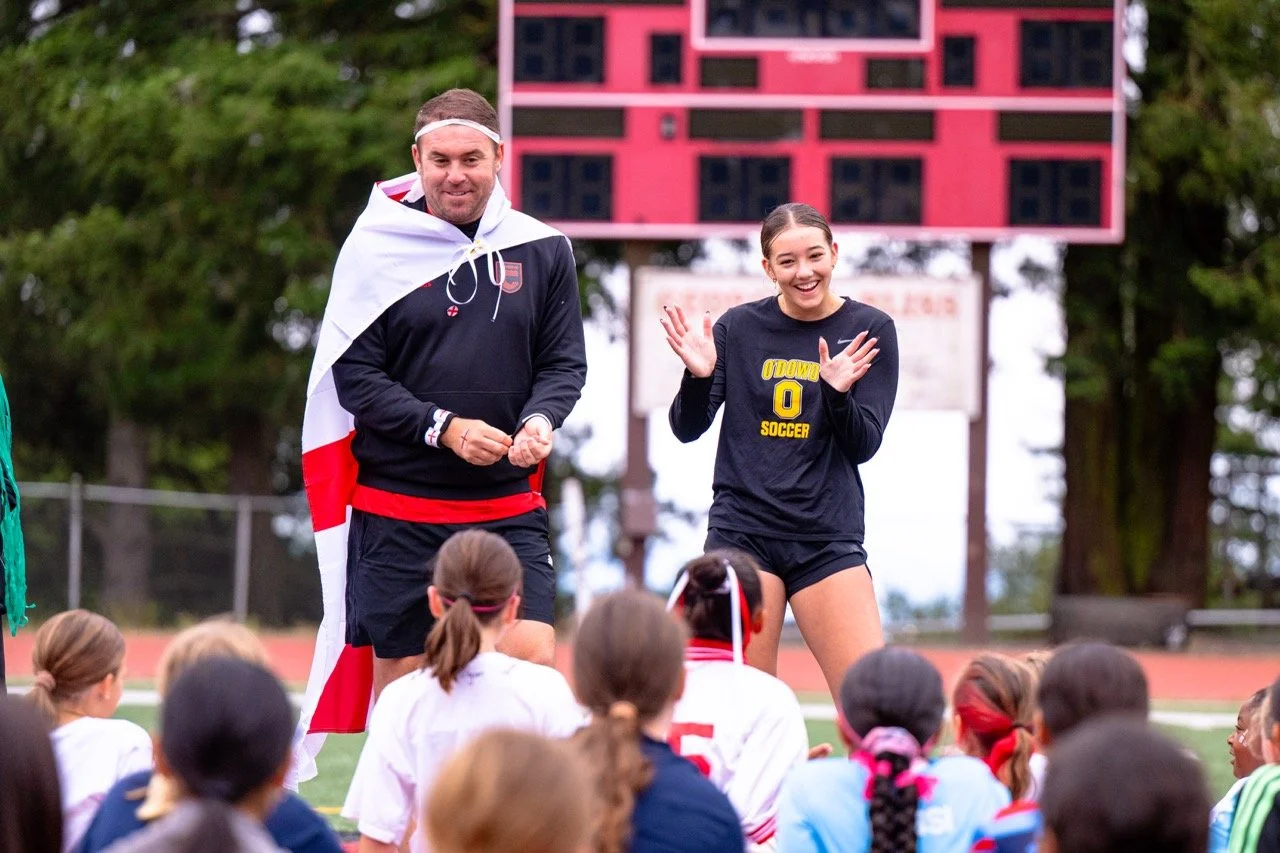 A man wearing a black sports jacket with a U.S. shield emblem and wrapped in a white and red flag, standing next to a smiling young woman in a black soccer uniform, surrounded by a group of young girls on a sports field.