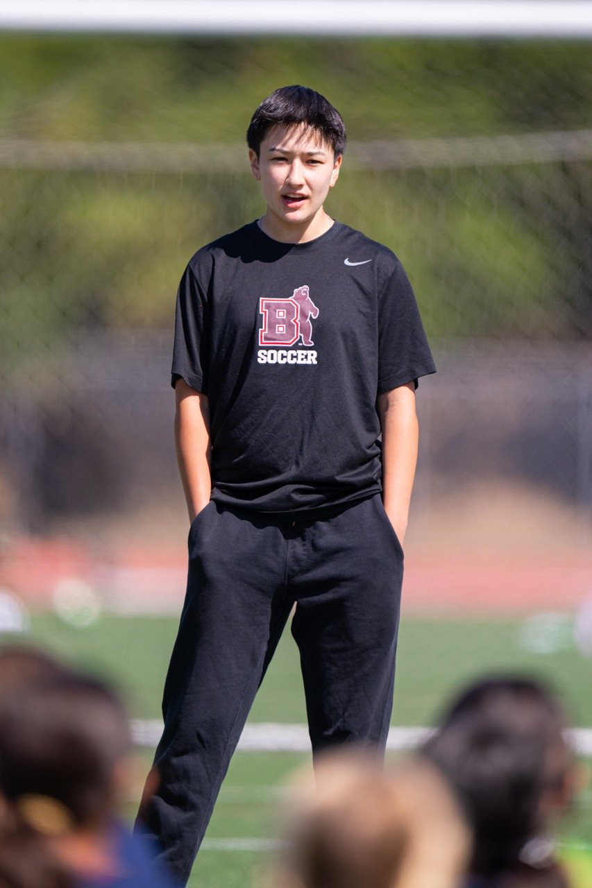 A young boy standing on a sports field, wearing a black soccer shirt and black pants, with hands in pockets and speaking to a group, with blurred spectators in the foreground.