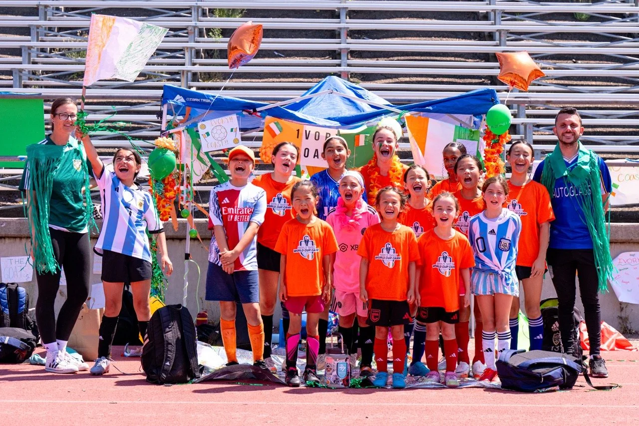 Group of young soccer players and coaches celebrating on a sports field with decorations and flags, some wearing orange and some in blue and white jerseys.