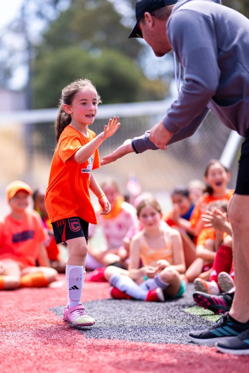 A young girl in an orange sports uniform high-fives a man during a sports event. Several children are sitting on a red track background, with some wearing orange shirts, and a chain-link fence and trees are visible in the background.