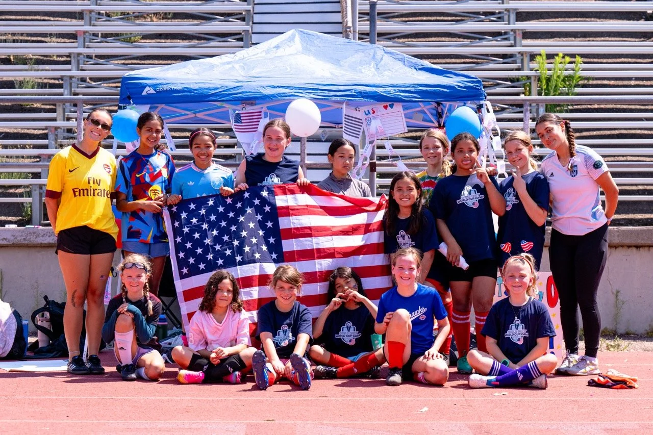 Group of young girls and women in sports uniforms, some holding an American flag, posing in front of a blue canopy at a sports stadium.