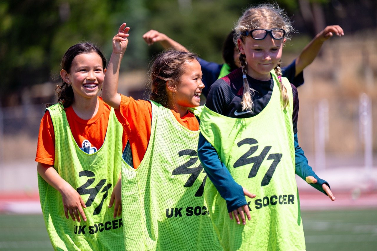 Group of young girls in yellow vests playing soccer on a field, smiling and enjoying the game.