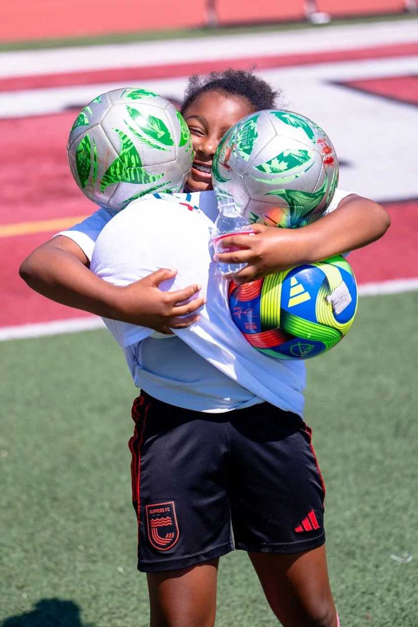 A young girl wearing black shorts and a white sports shirt joyfully hugging multiple soccer balls on a sports field.