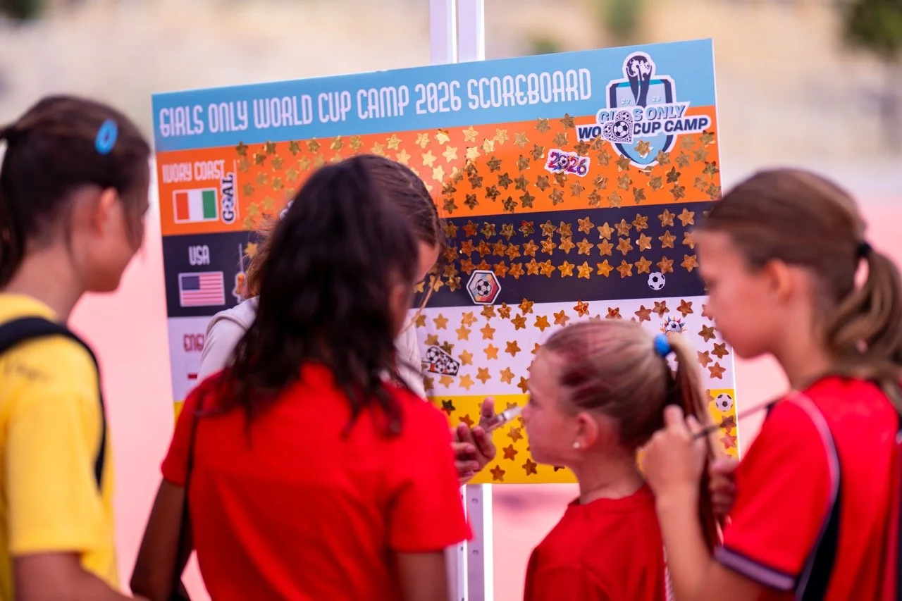 Young girls at a soccer camp looking at a large scoreboard poster titled 'Girls Only World Cup Camp 2026 Scoreboard' with flags and soccer-related graphics.