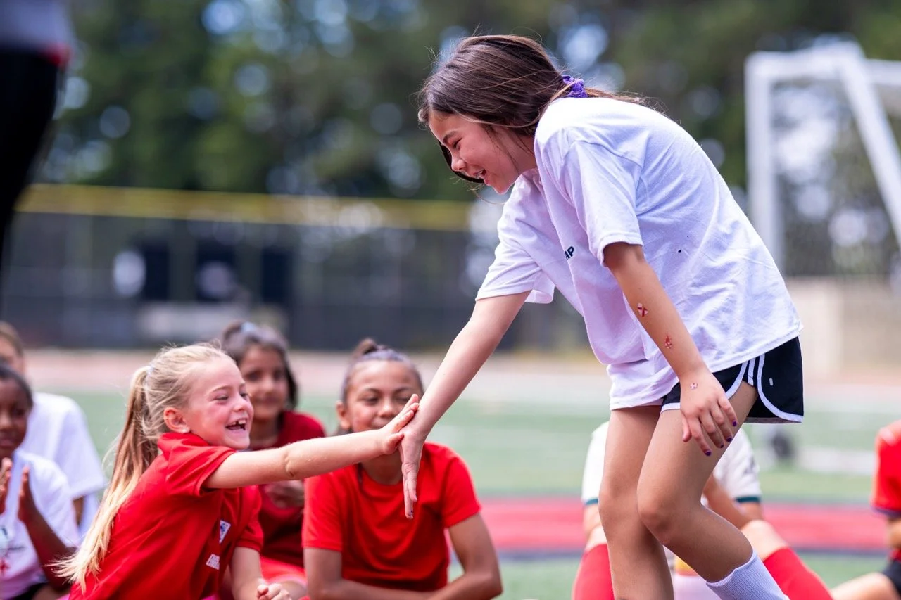 A young girl in a white t-shirt and shorts is playing a game with a group of girls in red shirts on a sports field. The girl in white is smiling and leaning forward, touching a girl's hand from the group who is also smiling, while others watch and sm