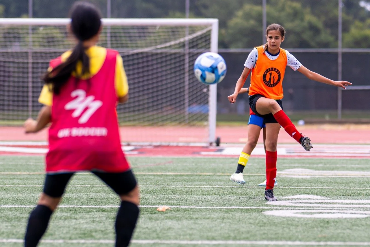 Two young girls playing soccer outdoors on a field, focusing on one girl wearing an orange bib and kicking a blue and white soccer ball, with a goal in the background, during daytime.